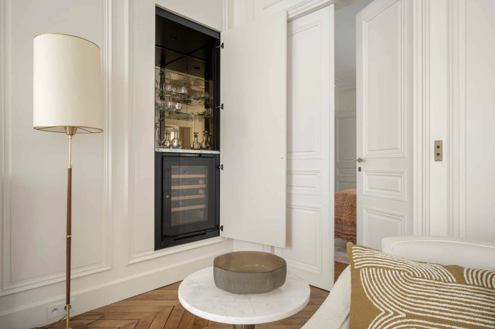 Living room corner with a tall floor lamp, a white wall with panel molding, a white door slightly open revealing a bed with red-orange bedding, a small circular marble-topped table with a brown bowl, and a recessed wall cabinet containing glassware a