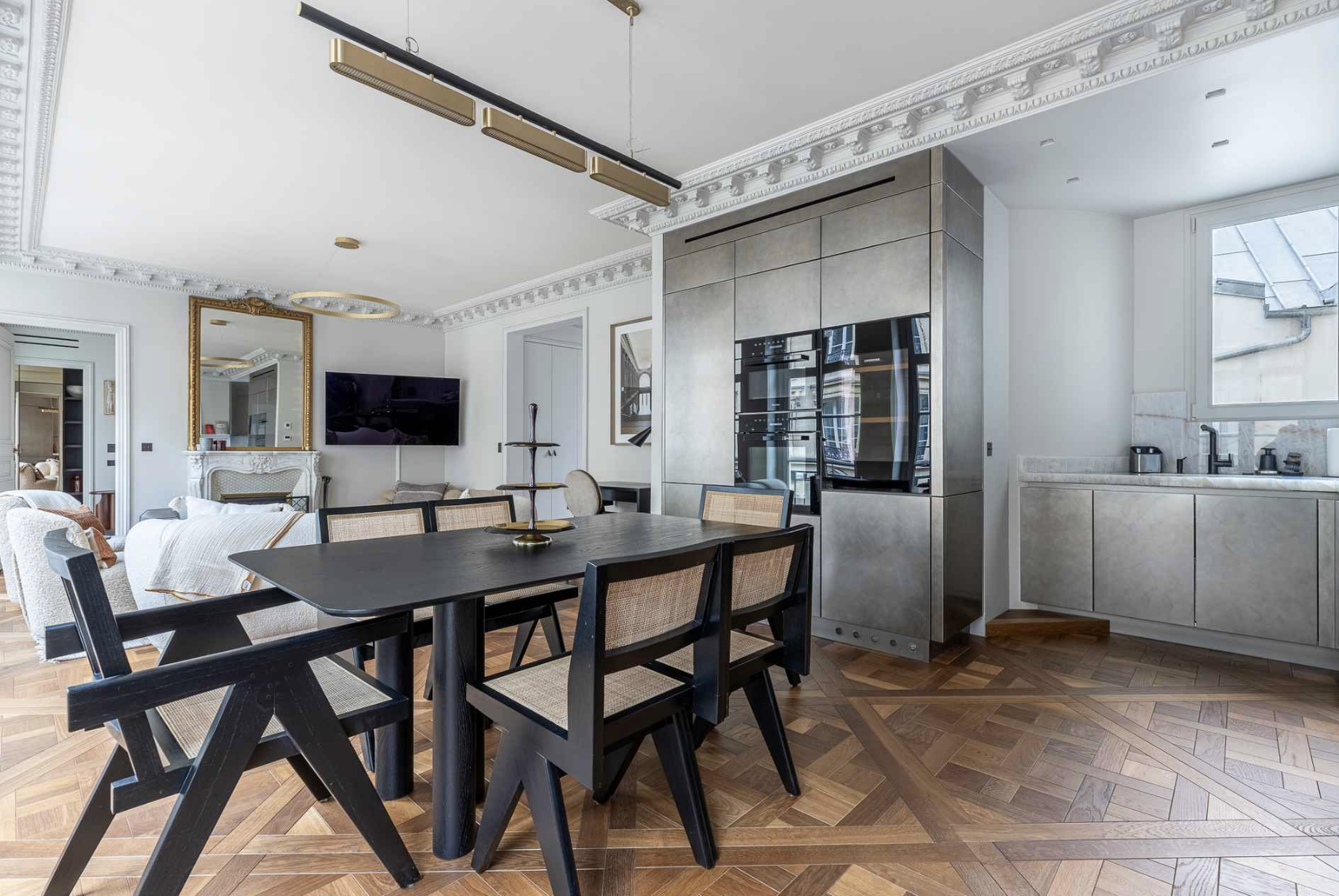 Modern kitchen and dining area with black dining table and chairs, gray cabinetry, and decorative crown molding on the ceiling, with a window over the sink and a television on the wall.