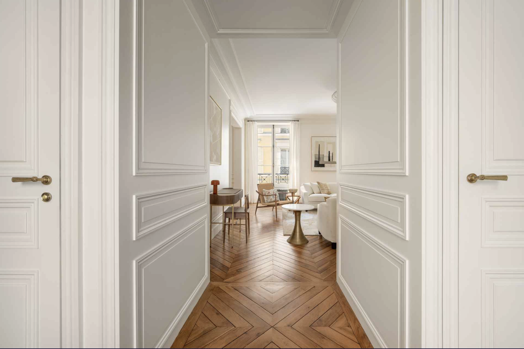View through a white double door into a bright living room with wooden herringbone floor, white walls, and art on the walls. There are chairs, a sofa, a small round table, and large windows with white curtains.