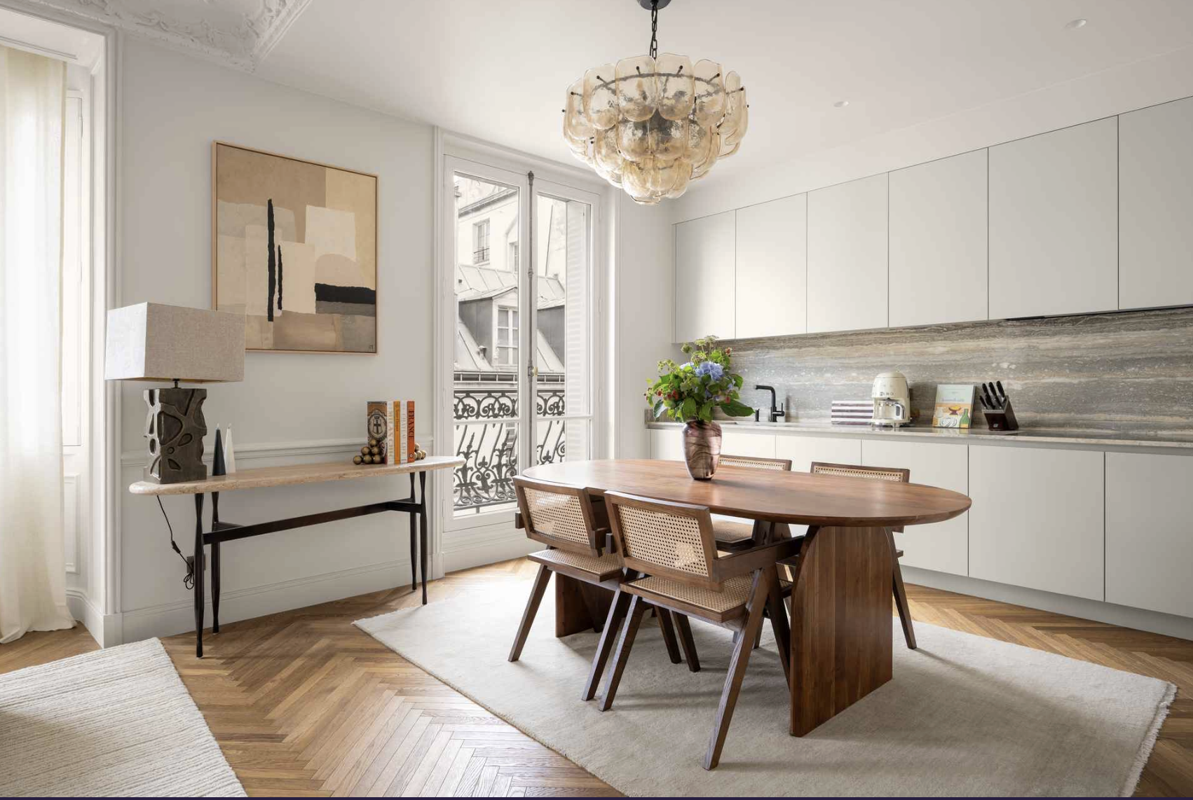 A dining area in a modern kitchen with a wooden oval table, four chairs, a vase with flowers, a chandelier, and a large window with a balcony view. There is a side table with decor and an abstract painting on the wall.