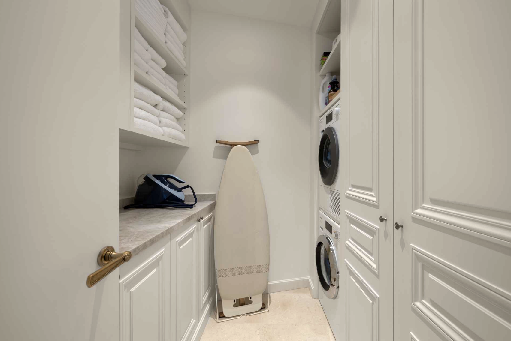 Laundry room with stacked washer and dryer, shelves with towels, ironing board, and an iron.