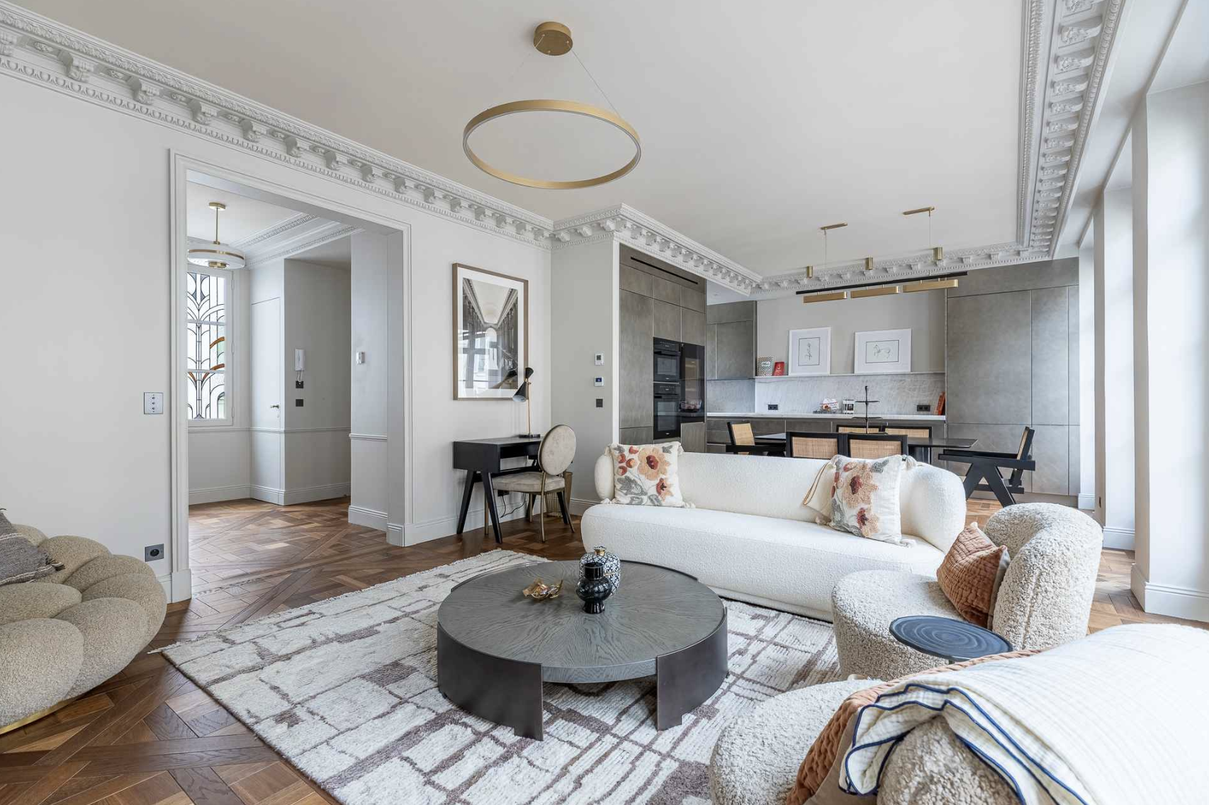 Modern living room with white sofas, patterned cushions, a round black coffee table, and a patterned rug, with an open kitchen in the background featuring gray cabinetry and minimalist decor.