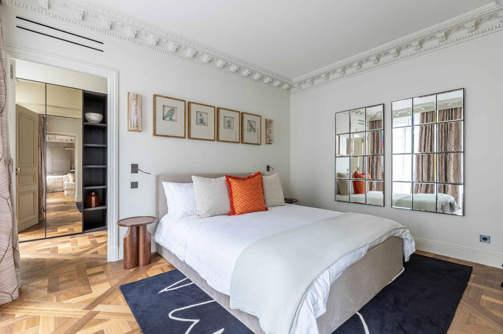 Bedroom with white walls, crown molding, a bed with white bedding and red accent pillow, wooden side table, black rug, and wall mirrors reflecting windows with curtains.