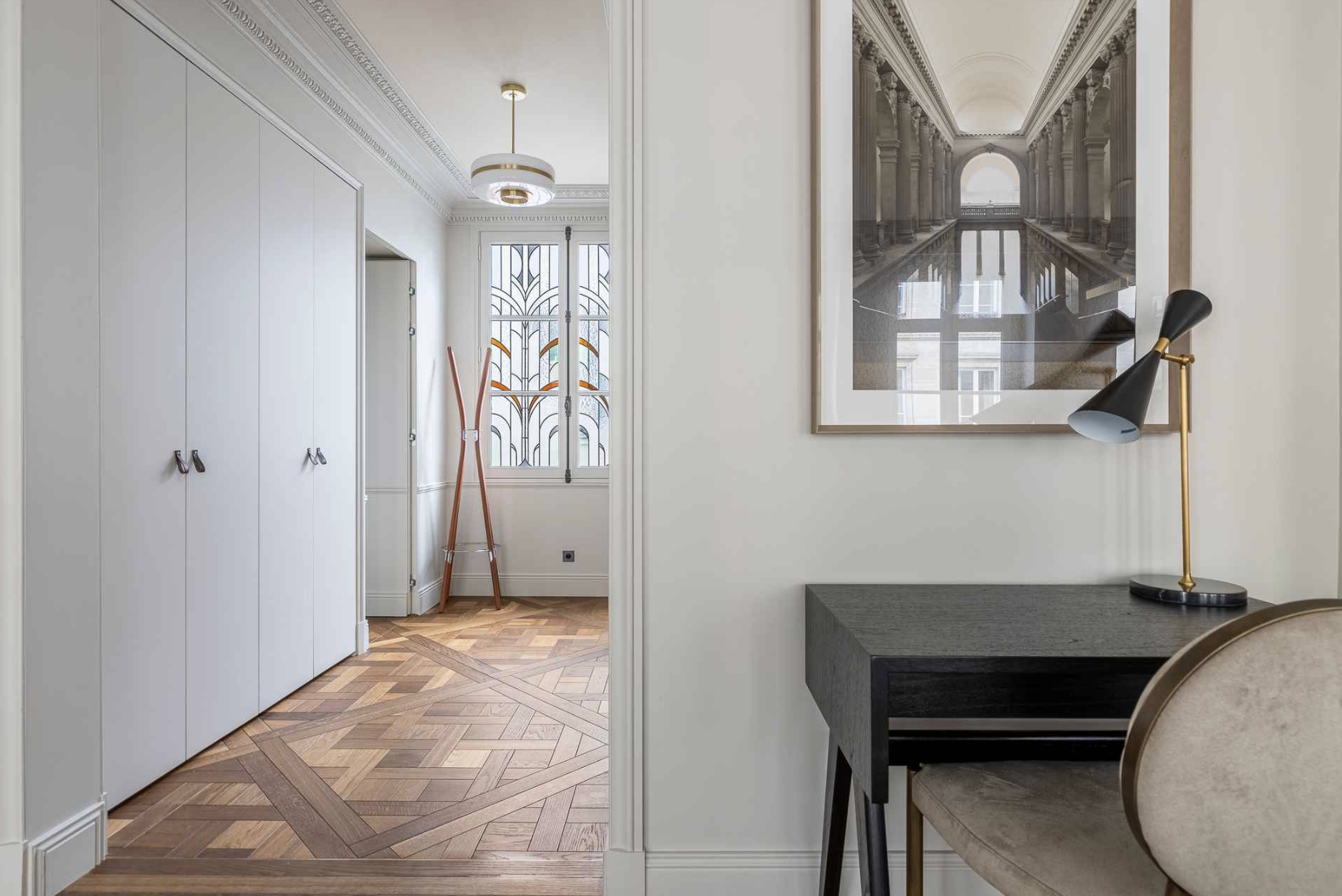 A view of a hallway with parquet wooden flooring, white walls, and a ceiling with decorative molding and a modern chandelier. There are large white closets on the left, a window with ornate ironwork, and a wooden coat rack. On the right, part of a ro