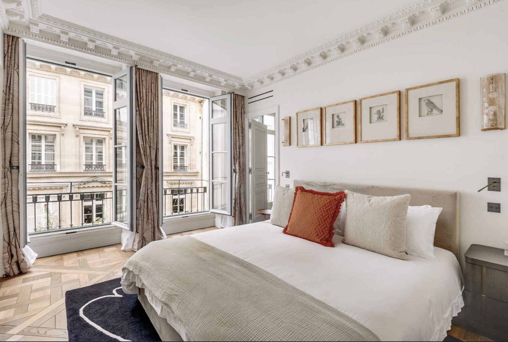 Bright bedroom with open French doors leading to a balcony, decorated with framed artwork and featuring a bed with white bedding and multiple pillows.