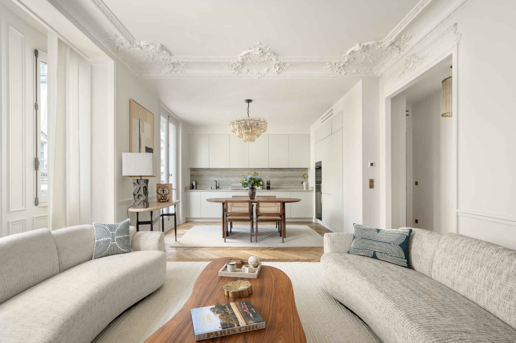 Open living room and kitchen with white walls, beige couches, a wooden coffee table with a book and decorative items, natural light from large windows, and a modern kitchen with white cabinets and a wooden dining table.