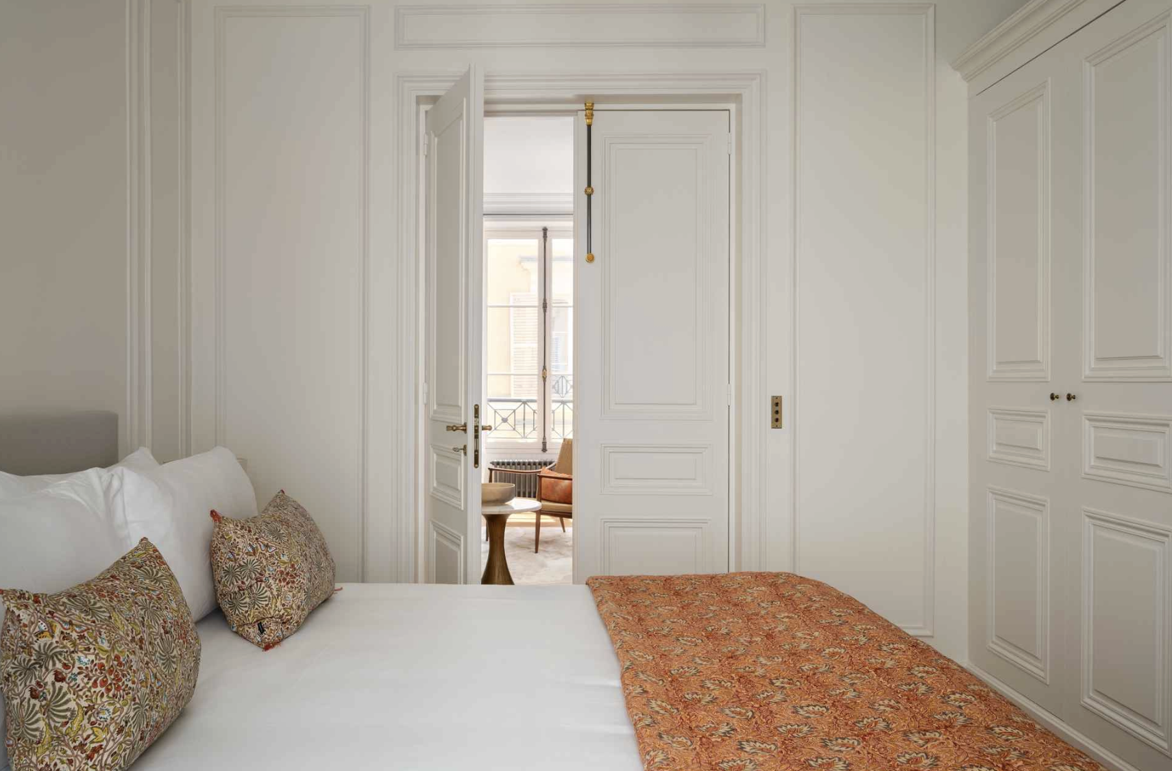 A bedroom with white walls and cabinetry, a bed with white bedding, patterned pillows, and an orange quilt, with a view of a sitting area through an open door leading to a balcony.