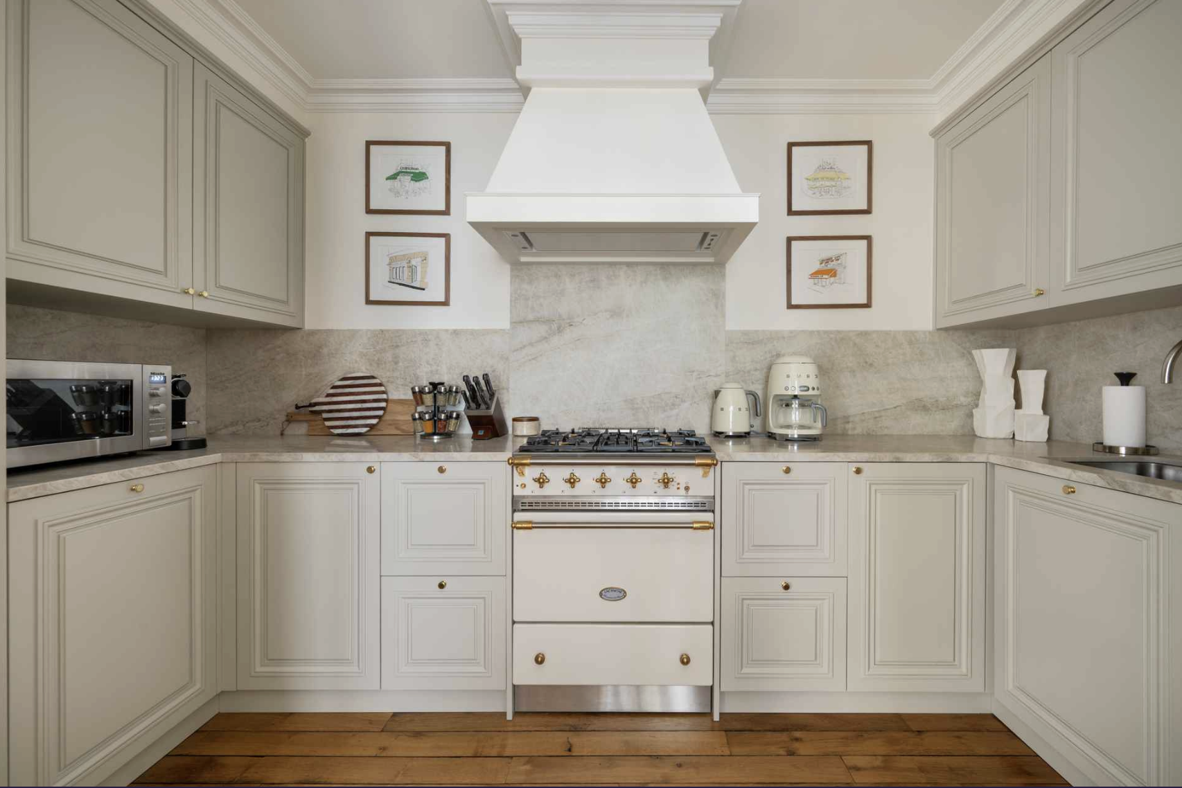 A modern kitchen with cream-colored cabinets, a stove with brass accents, and a marble backsplash. Kitchen appliances include a microwave oven, coffee maker, kettle, and toaster. Decor includes framed artwork and a wooden cutting board.
