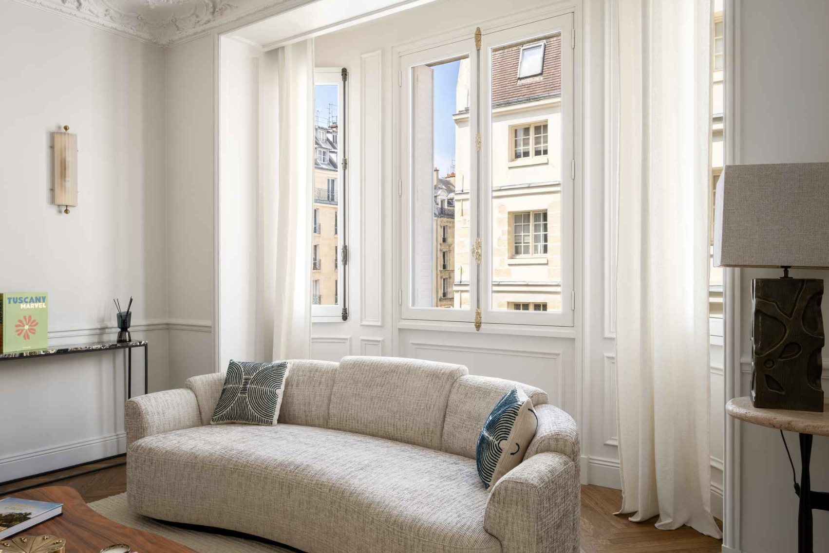 A bright living room with large windows, white walls, a beige sofa with patterned pillows, a table with a magazine, a floor lamp, and a small console table with a book and decor, overlooking neighboring buildings.