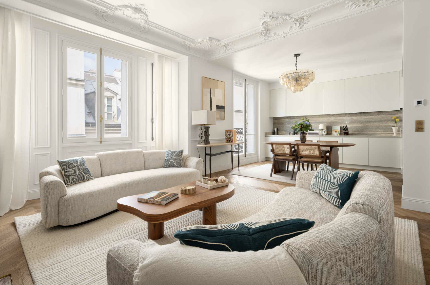 Living room with two beige sofas, wooden coffee table, and a dining area with a wooden table and four chairs, big windows with white curtains, and a modern chandelier ceiling light.
