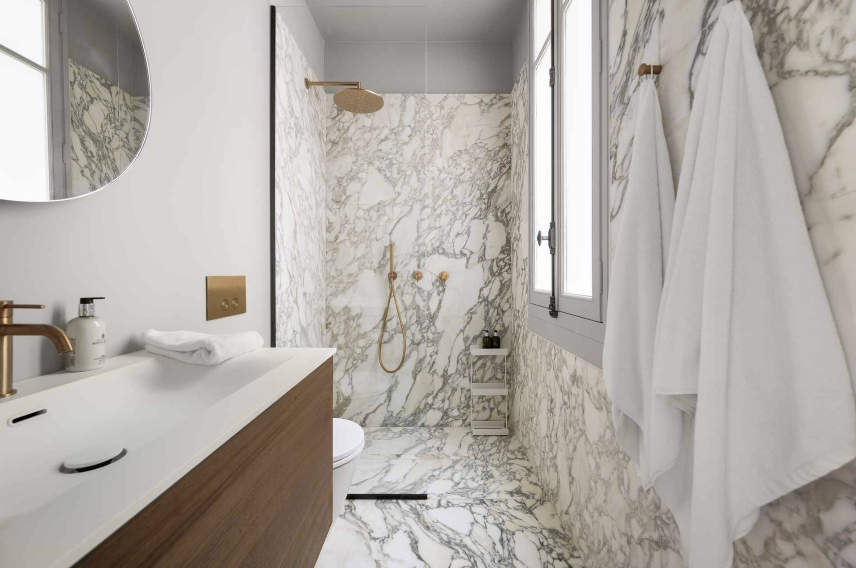 Modern bathroom with marble walls and floor, a window, a wall-mounted vanity with a white sink and gold fixtures, a mirror, a toilet, a shower with a rain shower head and gold fixtures, and white towels hanging on a wall hook.