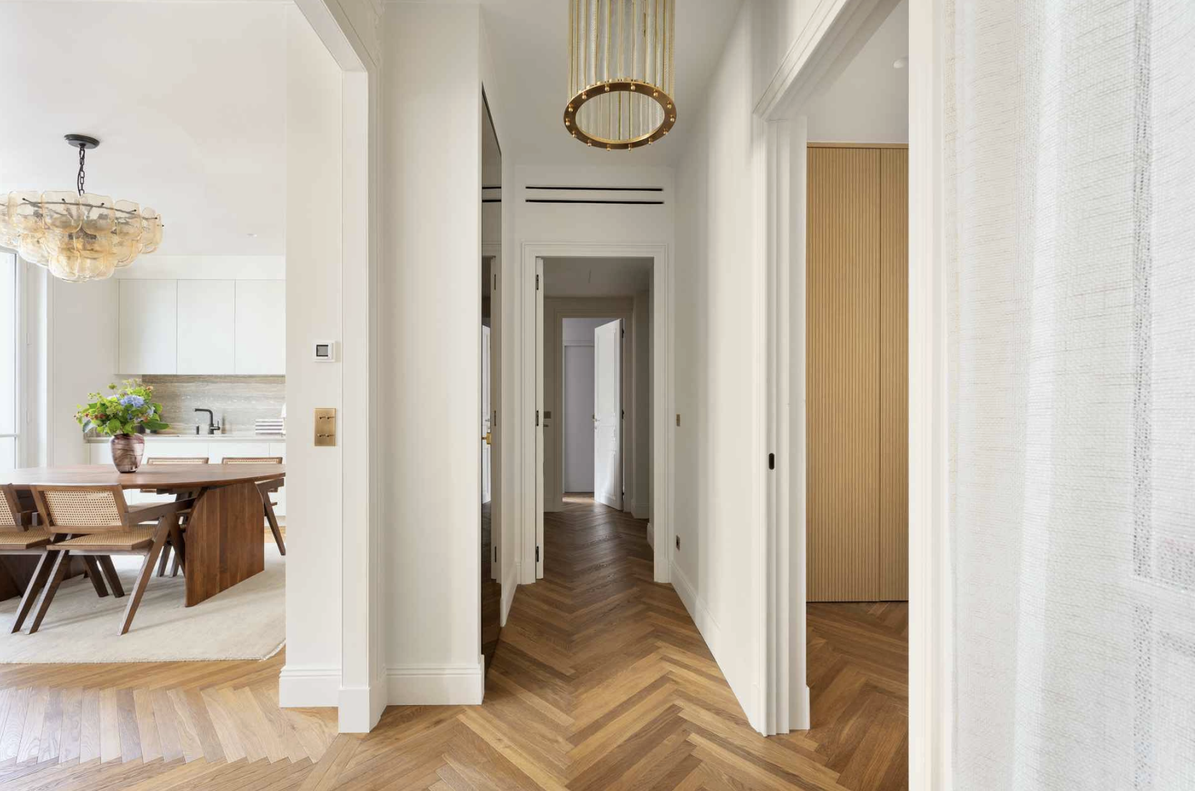 Hallway with wooden herringbone floor, white walls, and doors leading to various rooms, with modern ceiling light fixtures.