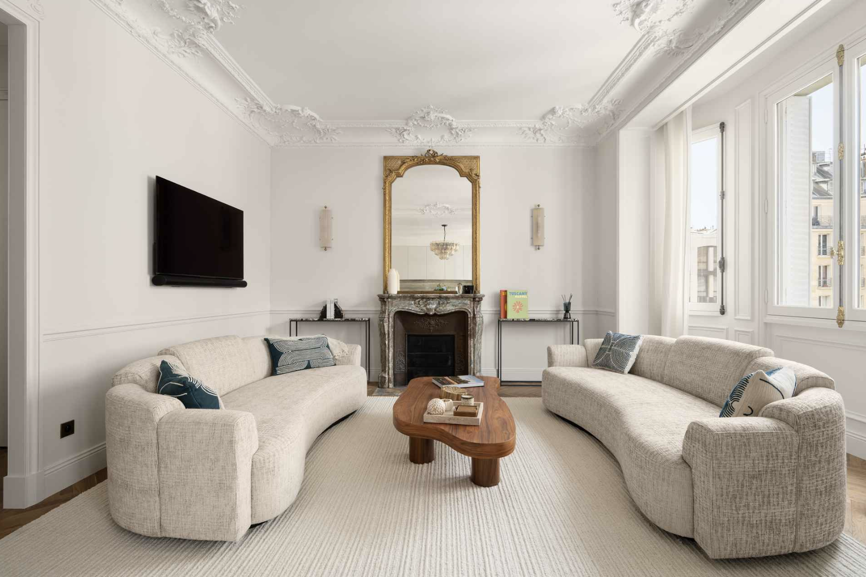 Living room with two beige curved sofas, a wooden coffee table with books, a fireplace with a mirror above, and a flat-screen TV mounted on the wall. Large windows with white curtains let in natural light.