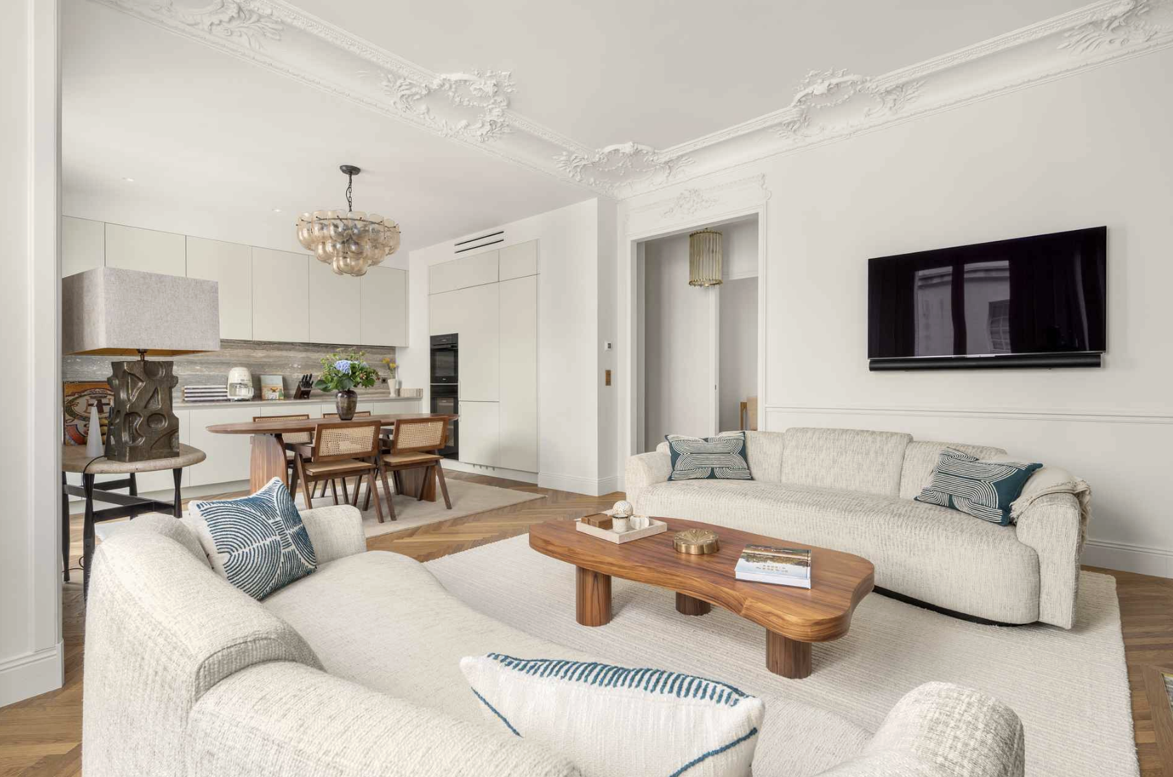 Living room with two off-white sofas, a wooden coffee table, and a TV mounted on the wall. The room features ornate white ceiling moldings and hardwood floors. In the background, an open kitchen with white cabinets, a dining table, and chairs.