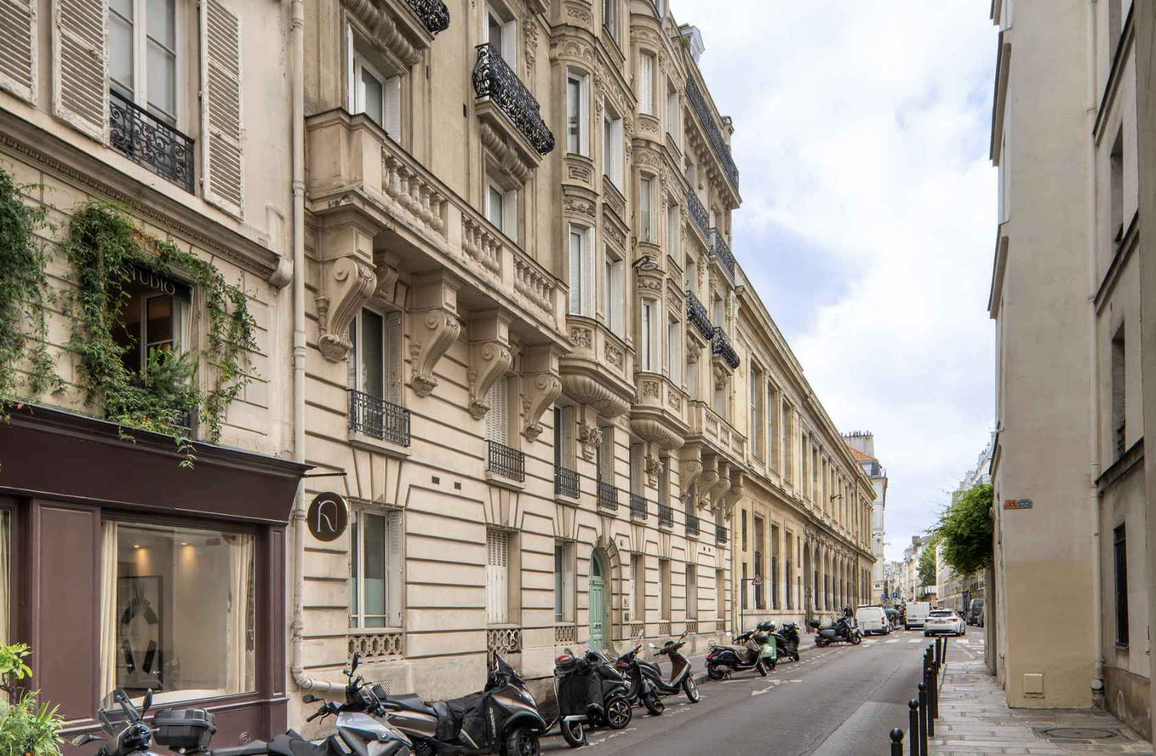 Paris street scene with classical beige buildings, wrought iron balconies, parked scooters, and cars along the curb under a cloudy sky.