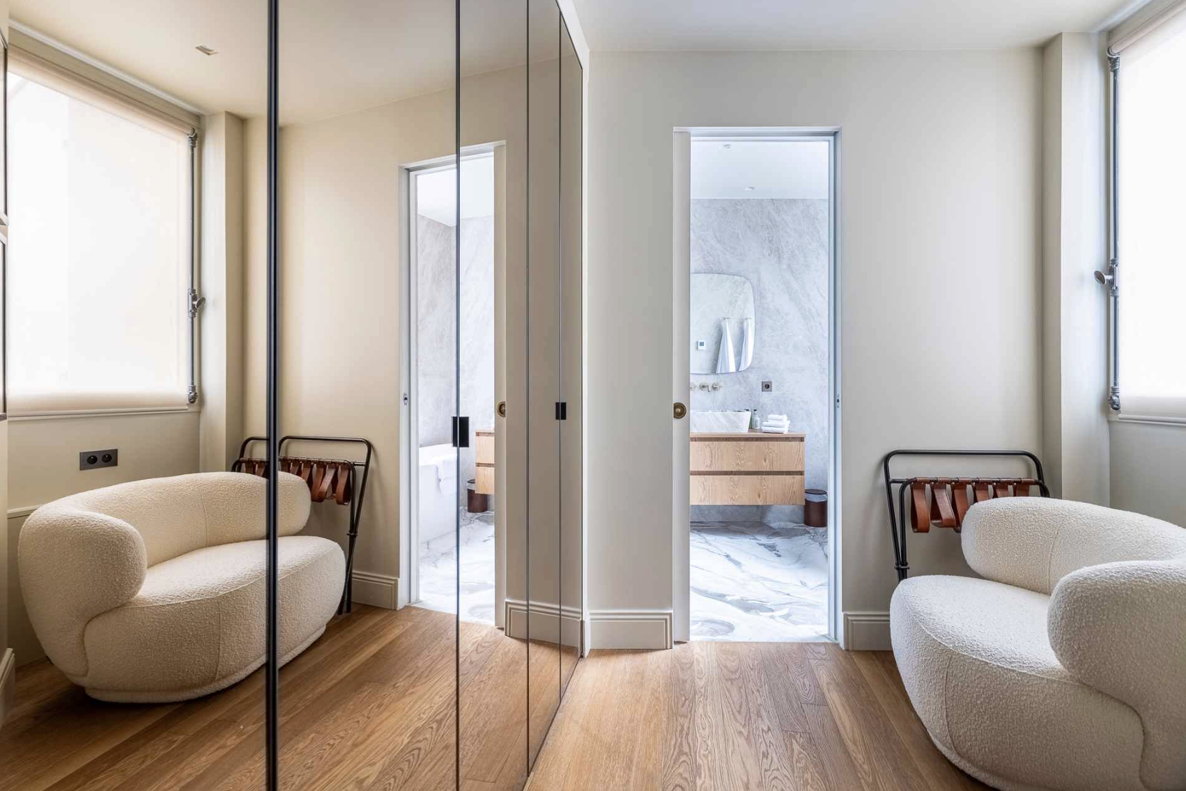 Interior of a modern bedroom with two white textured armchairs, a mirrored wardrobe, and an en-suite bathroom with marble floors and wooden vanity.