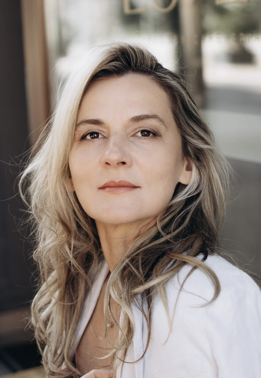 A woman with long wavy blonde hair, wearing a white top, looking confidently into the camera, seated at a cafe or restaurant with a blurred background.