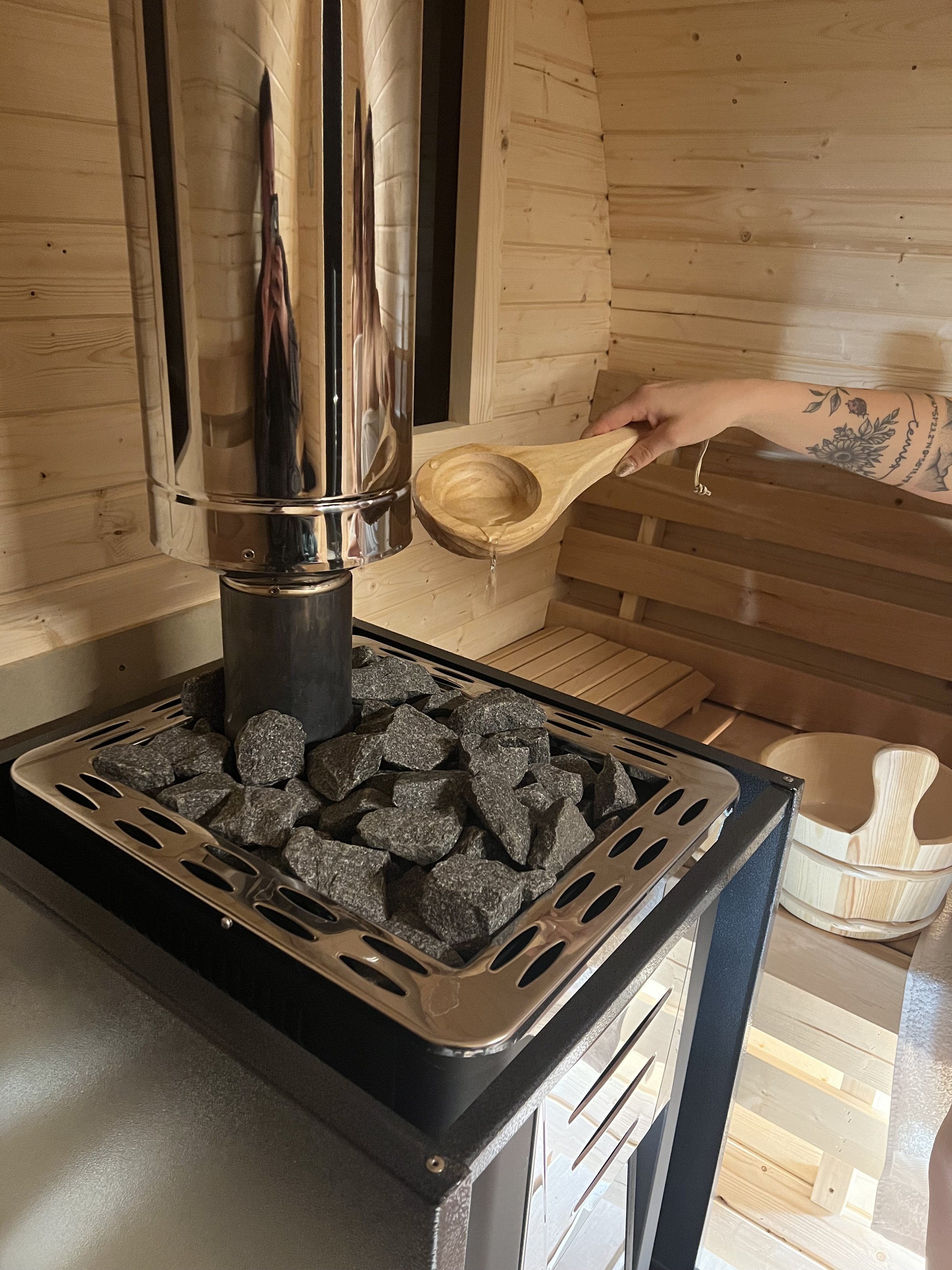 Person gießt Wasser in einen Holzlöffel, neben einem Saunaofen mit Steinen in einem Holzraum.