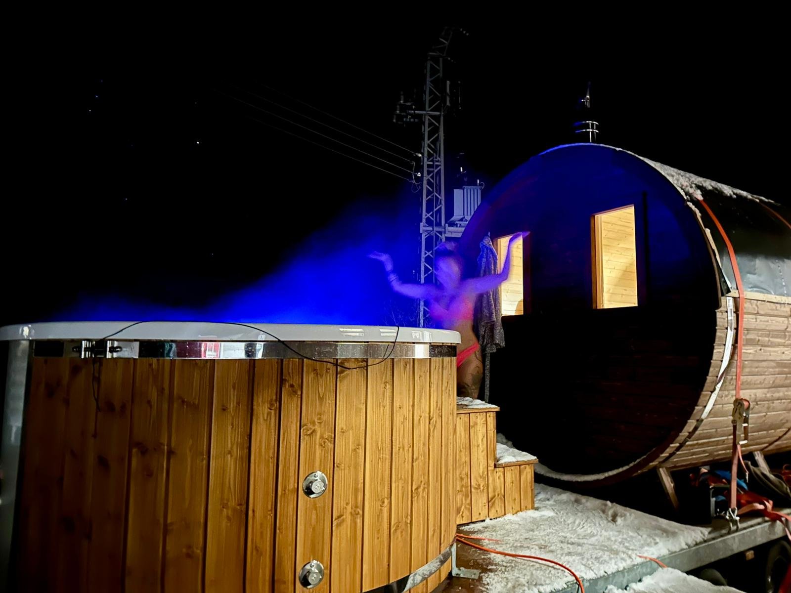 Person in Badekleidung beim Ausstieg aus einer Holzhütte auf Schnee bei Nacht, mit heißem Dampfausstoß, umgeben von Holz und Eiszapfen, dunkler Himmel im Hintergrund.