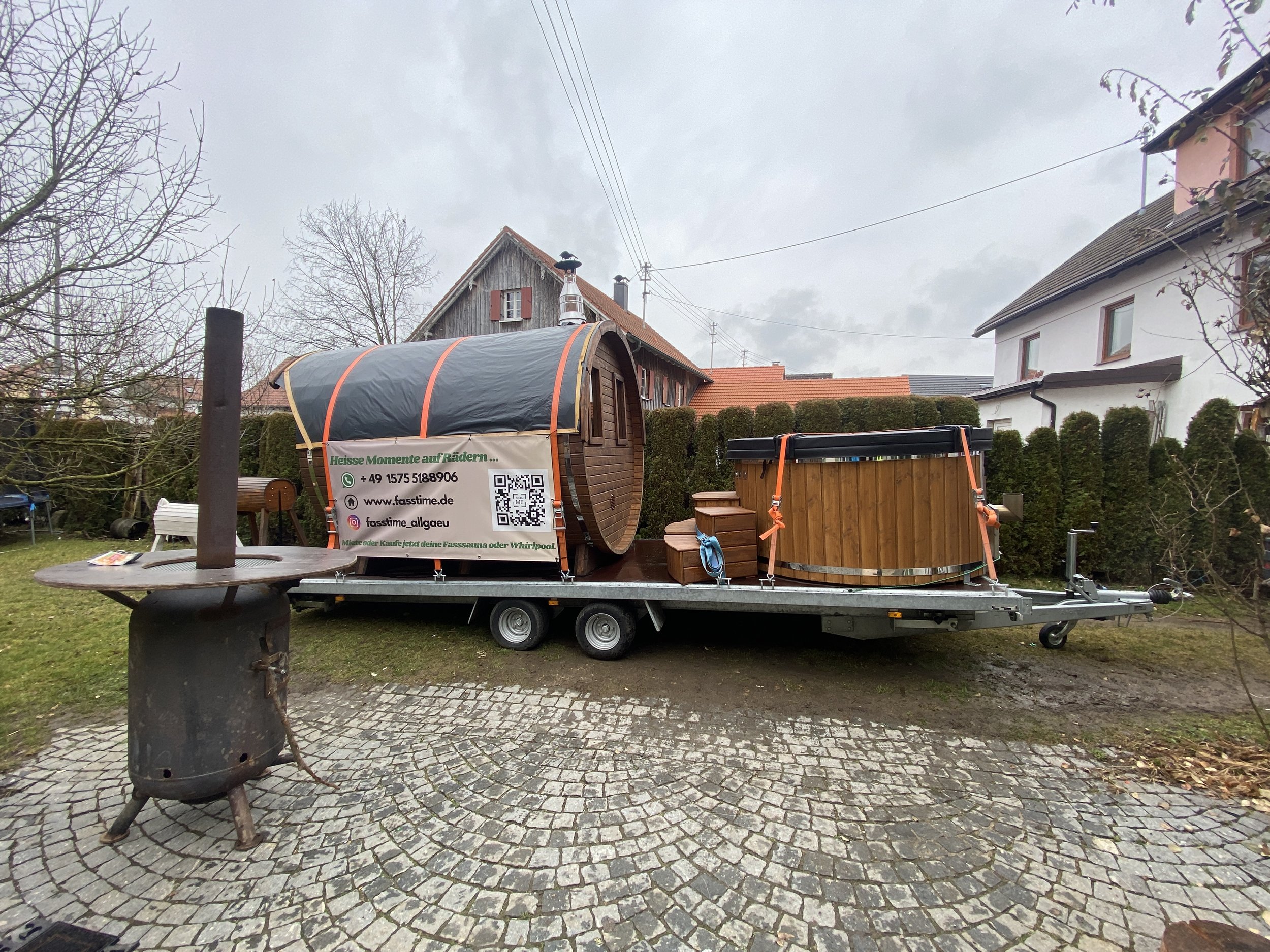 Ein Wohnwagen mit einem Saunaanhänger auf einem Anhänger beladen, im Freien auf einem gepflasterten Bereich, umgeben von Bäumen und Häusern. Es gibt einen Holzofen im Vordergrund.