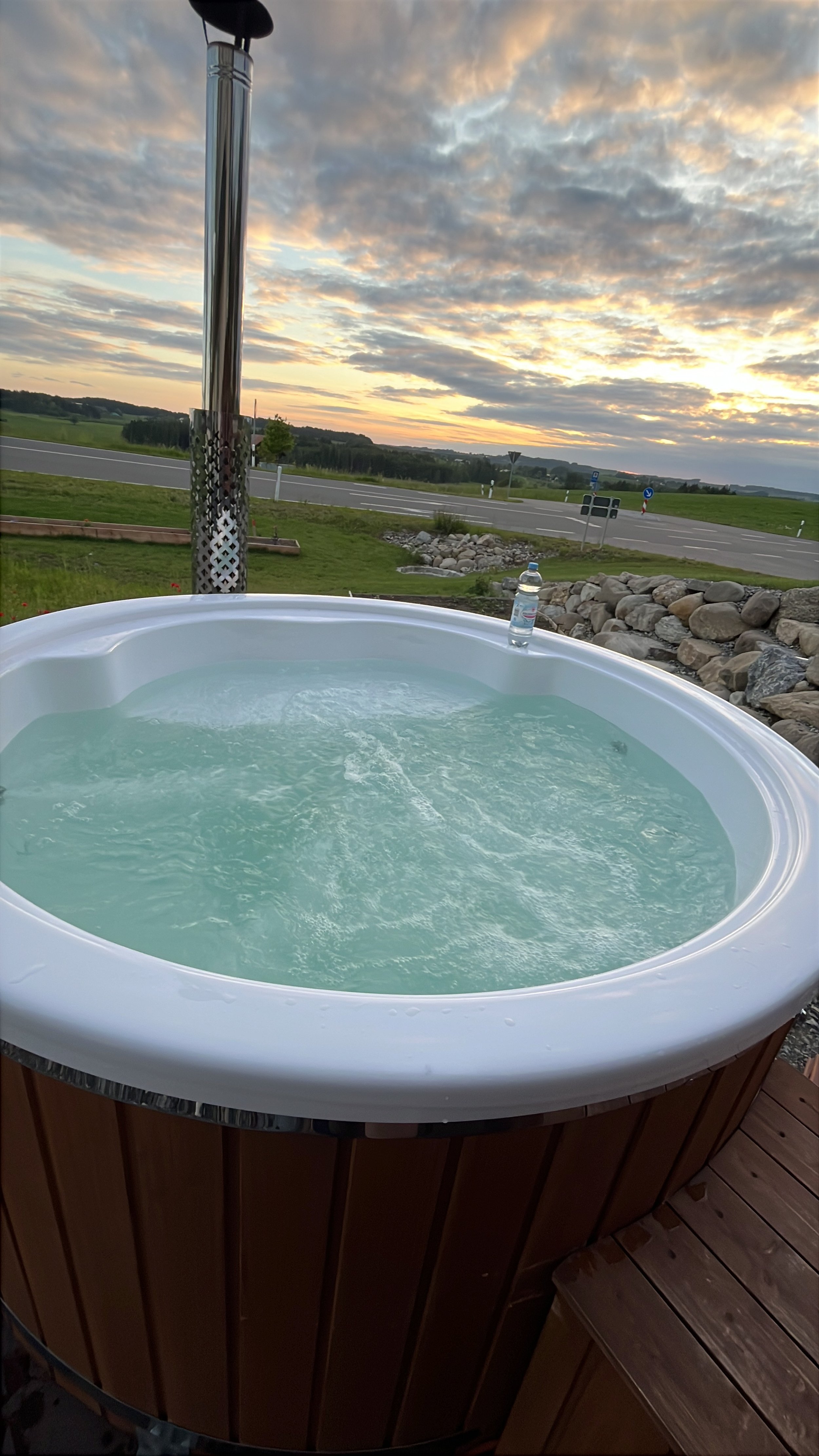 Ein Whirlpool im Freien mit Blick auf einen Sonnenuntergang, Wasserflasche auf der Kante des Whirlpools und eine offene Landschaft im Hintergrund.
