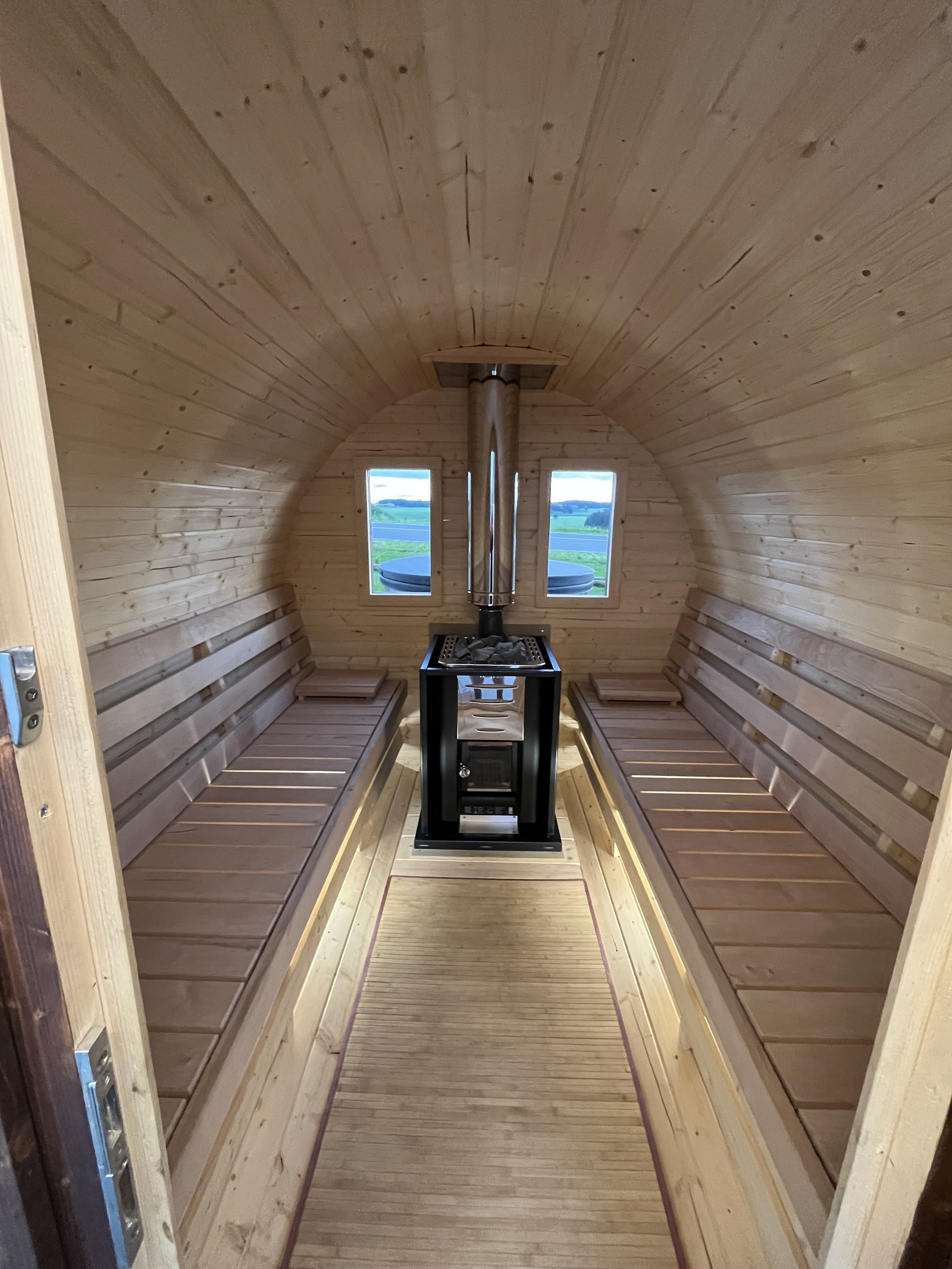Innenraum einer Saunakabine mit Holzsitzen, Holzdecke, einem Kamin in der Mitte und zwei kleine Fenster