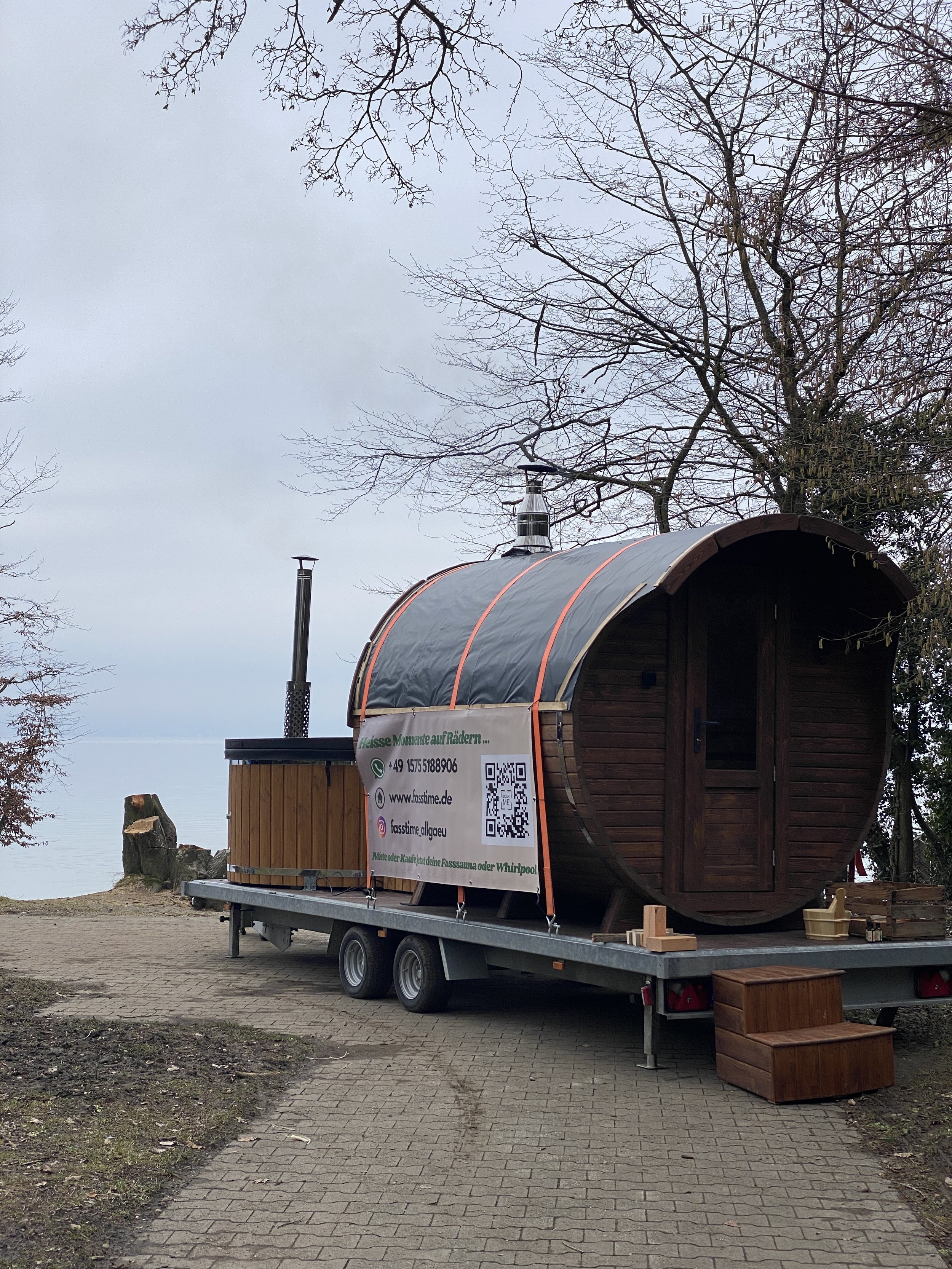 Ein Holzsauna auf einem Anhänger, eingebettet in eine natürliche Umgebung mit Bäumen, vor einem See bei bewölktem Himmel.