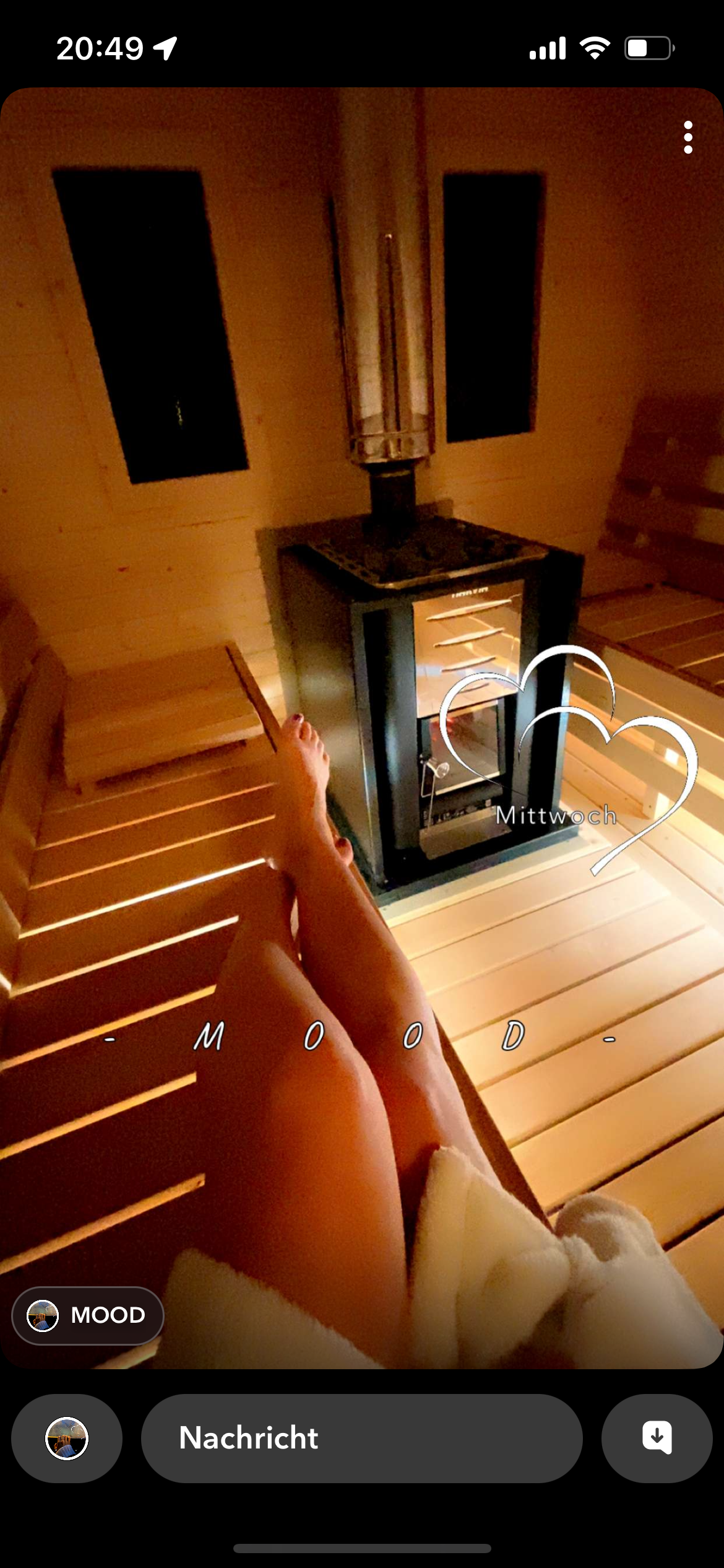 Innenaufnahme einer Sauna mit Holzbank, eine Person liegt entspannt mit hochgelegten, nackten Beinen, im Hintergrund ein Saunaofen