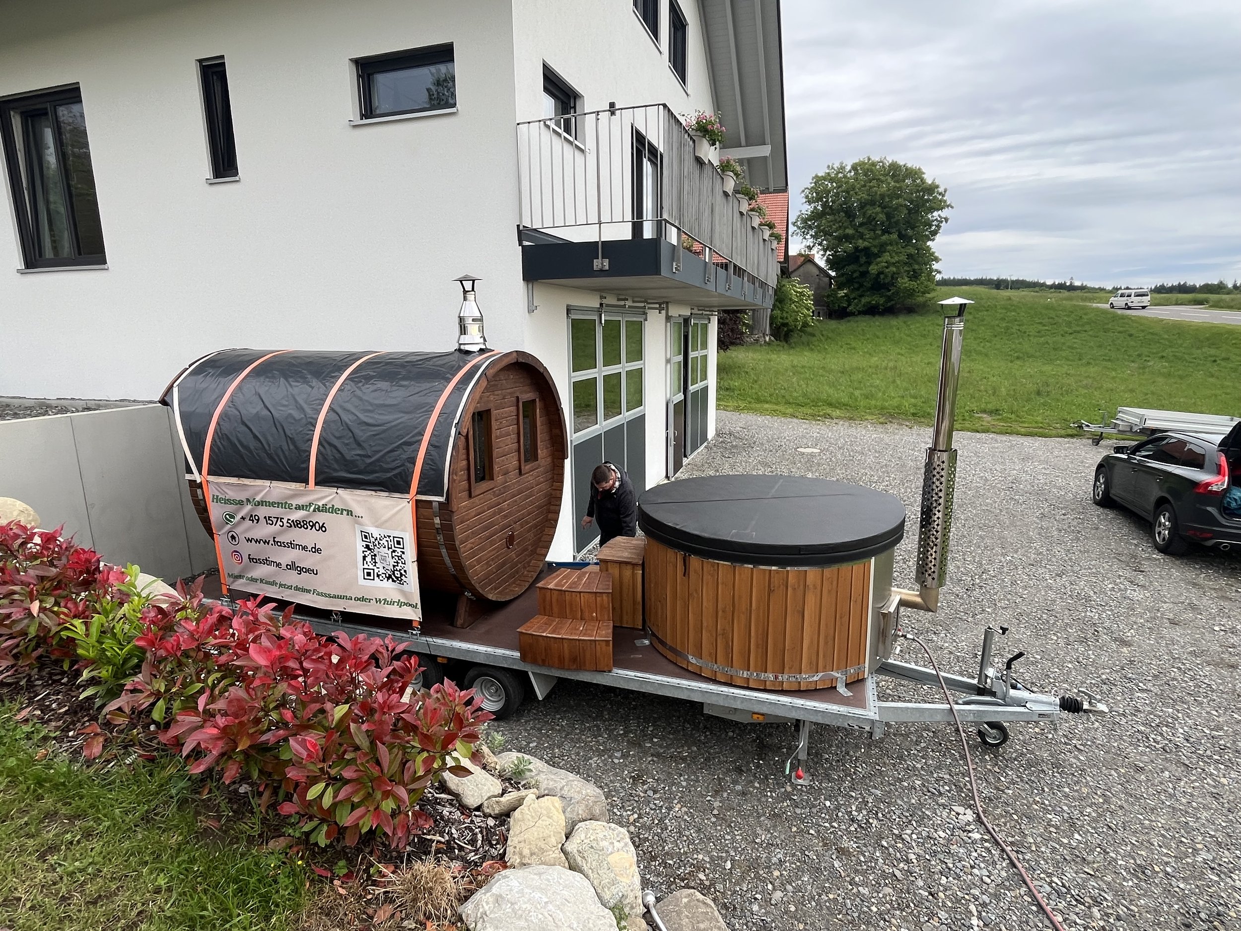 Holzzuber für Saunagänge auf Ladefläche vor modernem Mehrfamilienhaus, mit einer Person am Eingang, umgeben von Grünflächen und Parkplätzen.