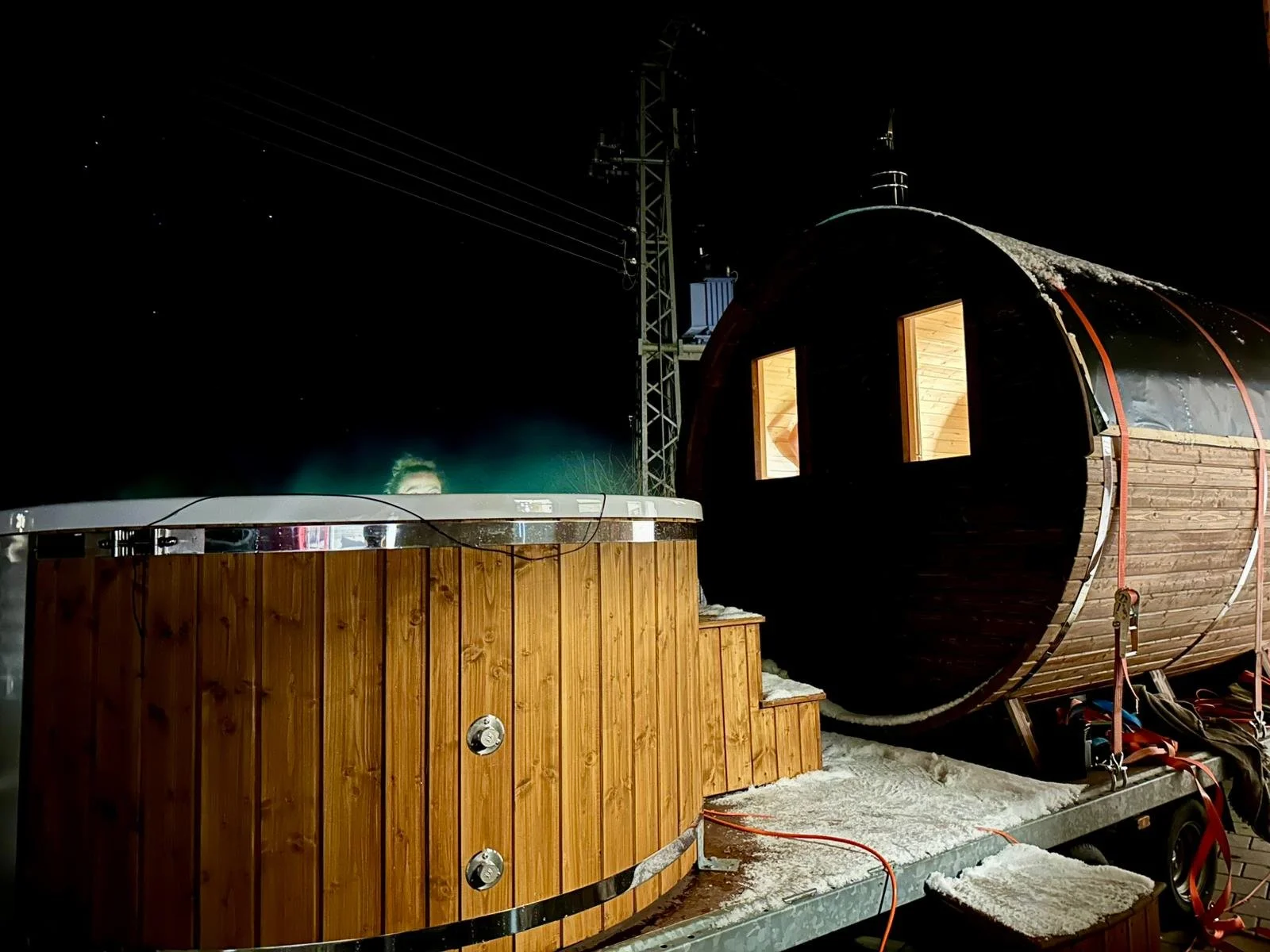 Holzhaus am Wasser mit Hot Tub bei Nacht, umgeben von Schnee, mit Strommast im Hintergrund.