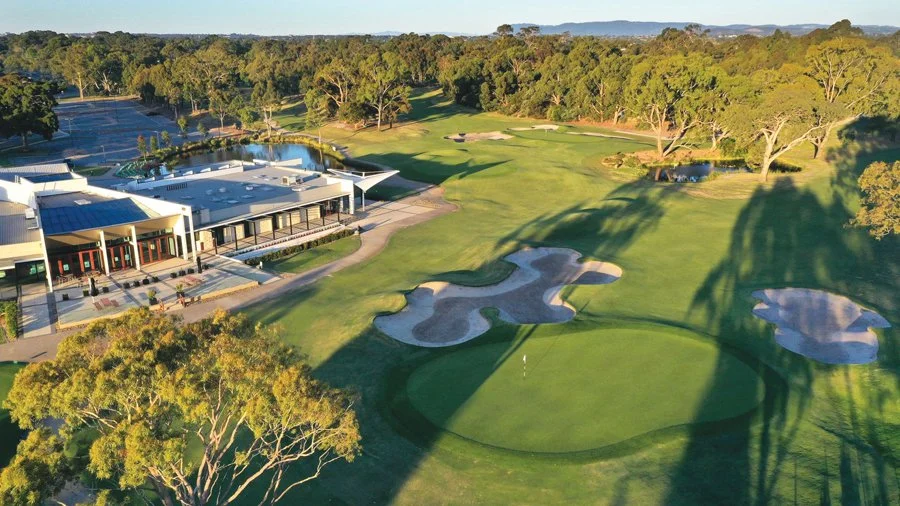Aerial view of a golf course with green, sand bunkers, trees, and a clubhouse, during a sunny day.