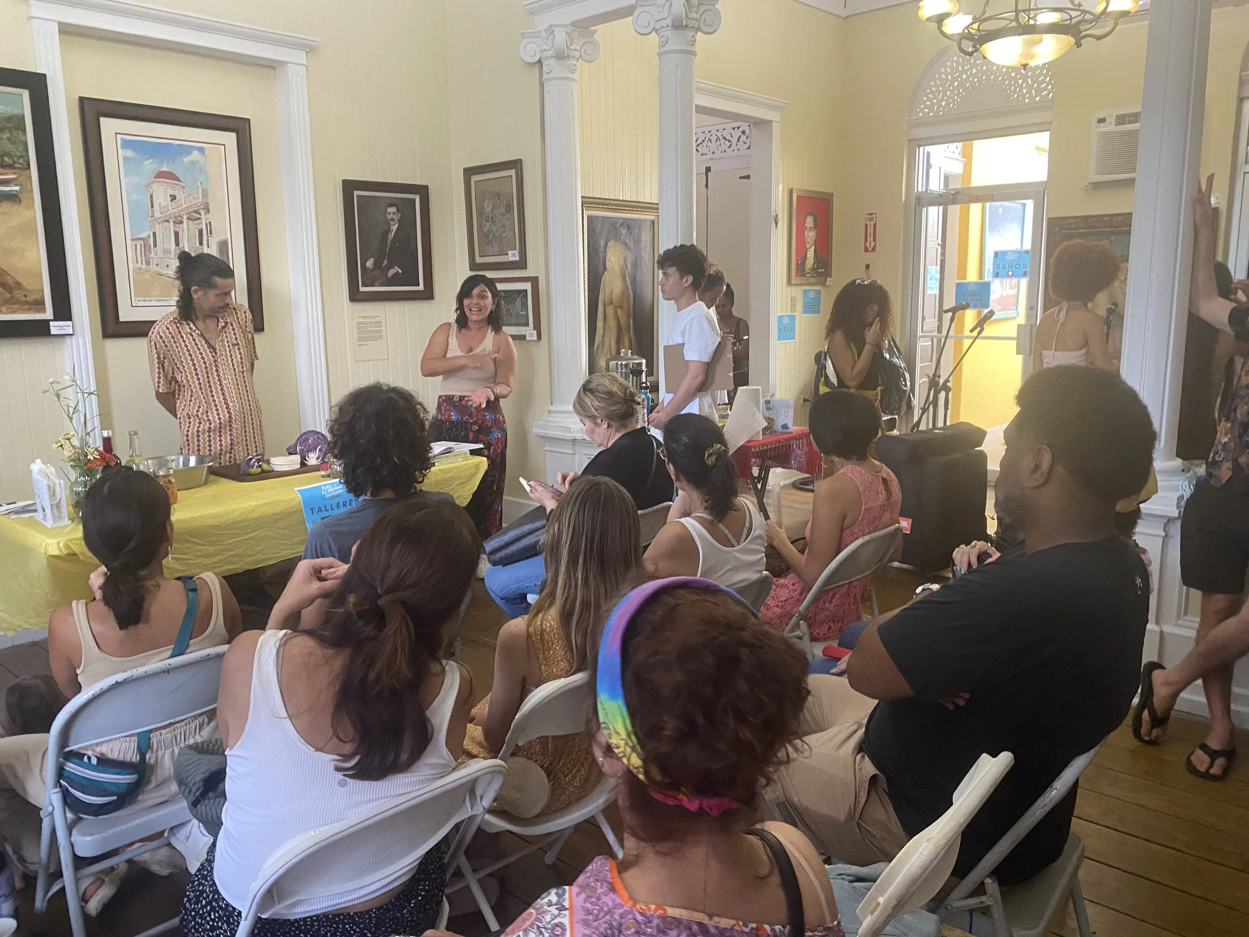 Varios asistentes en una exposición de arte en un espacio con paredes amarillas, decoradas con cuadros y retratos. Un grupo de personas está sentado en sillas blancas y otras de pie, conversando y viendo la exposición. En el centro, una mujer con blusa sin mangas y falda florada parece estar hablando o dando una presentación.