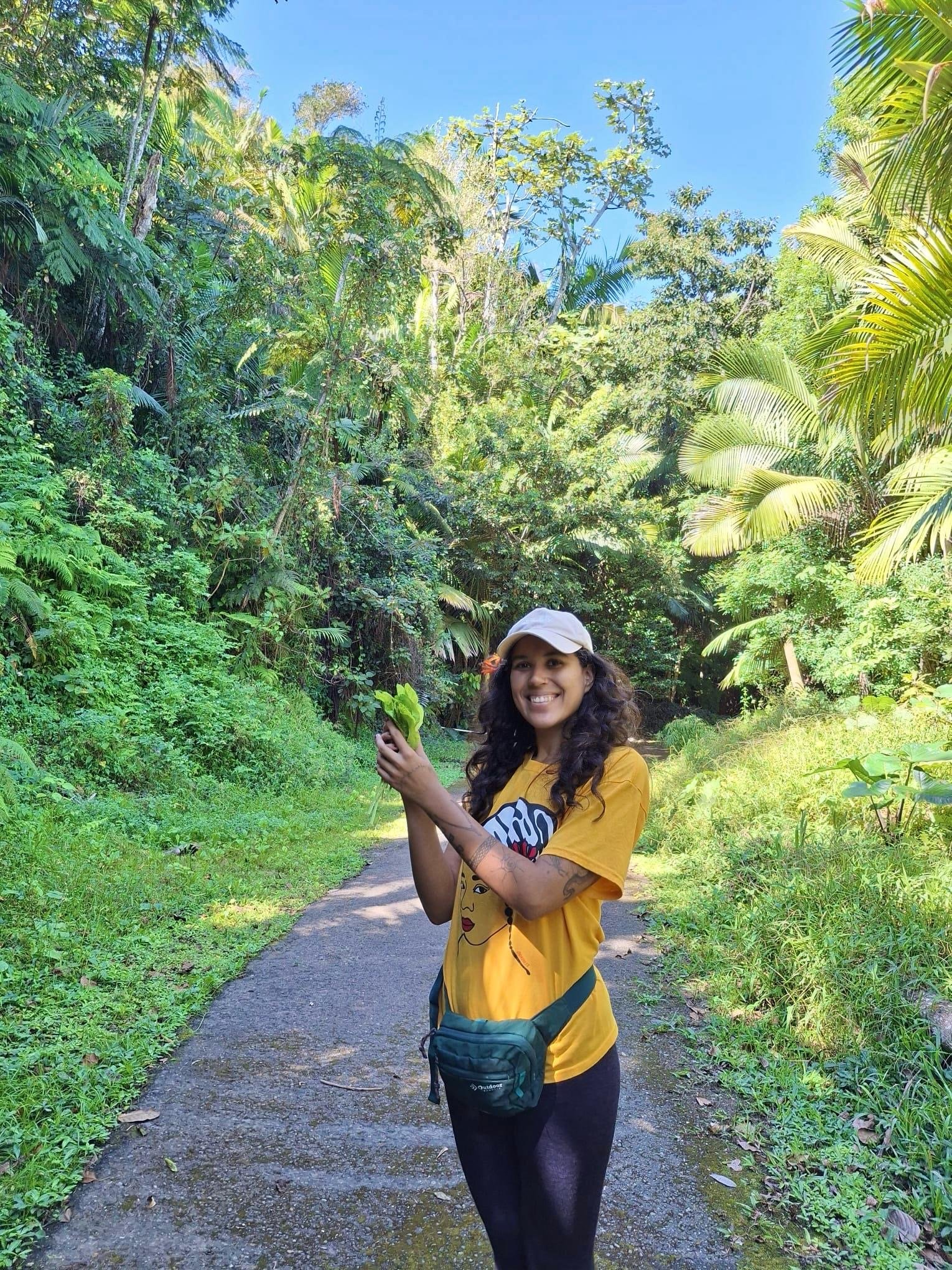 Mujer sonriendo en un sendero rodeada de vegetación densa en un entorno natural, sostiene una hoja verde y lleva puesta una gorra blanca, camiseta amarilla y una bolsa de cadera.