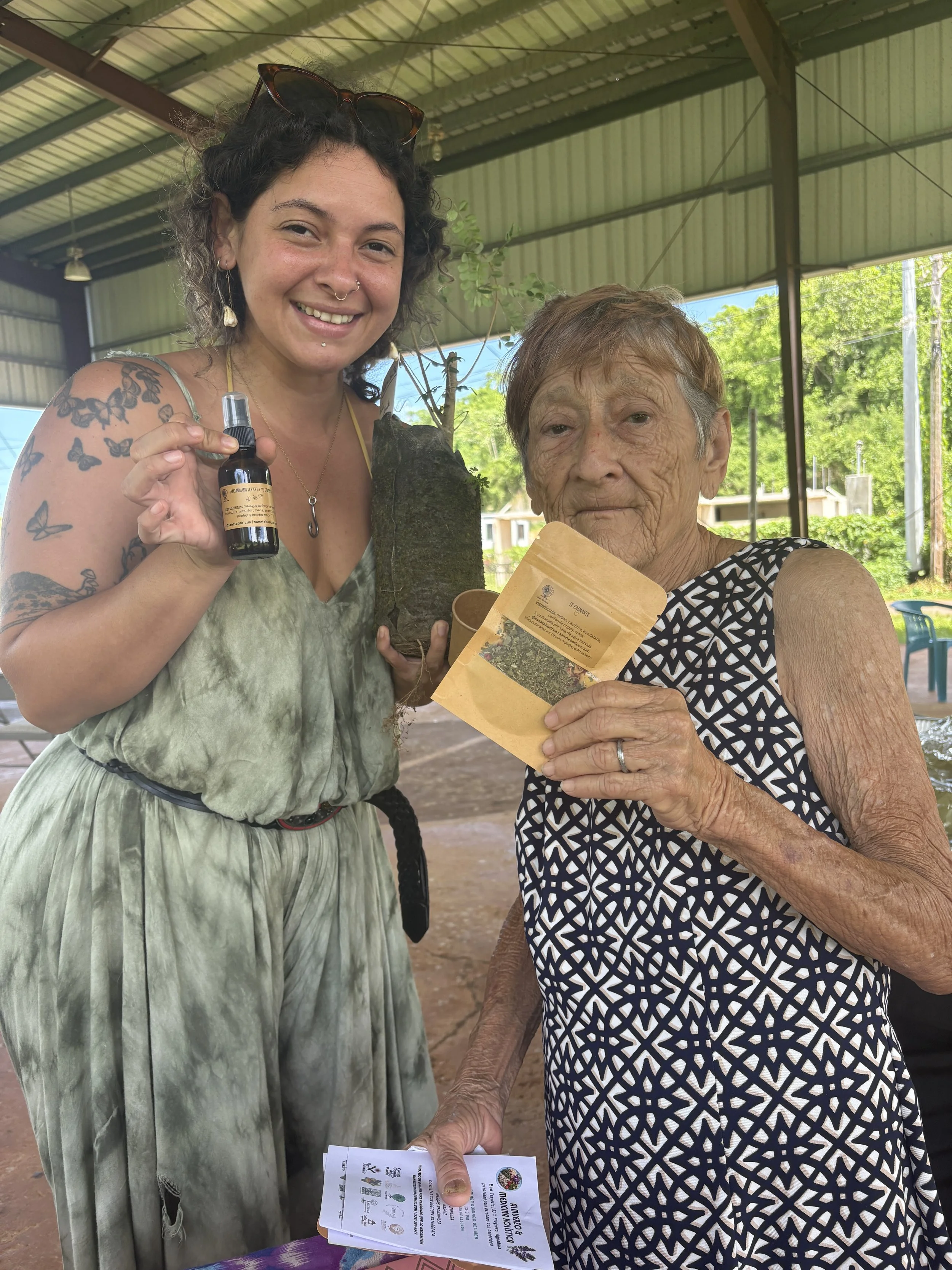 Una mujer joven con tatuajes y accesorios, sosteniendo un frasco, junto a una mujer mayor con gafas, ambas sosteniendo documentos y una planta, en un espacio abierto con estructura metálica y fondo de árboles verdes.