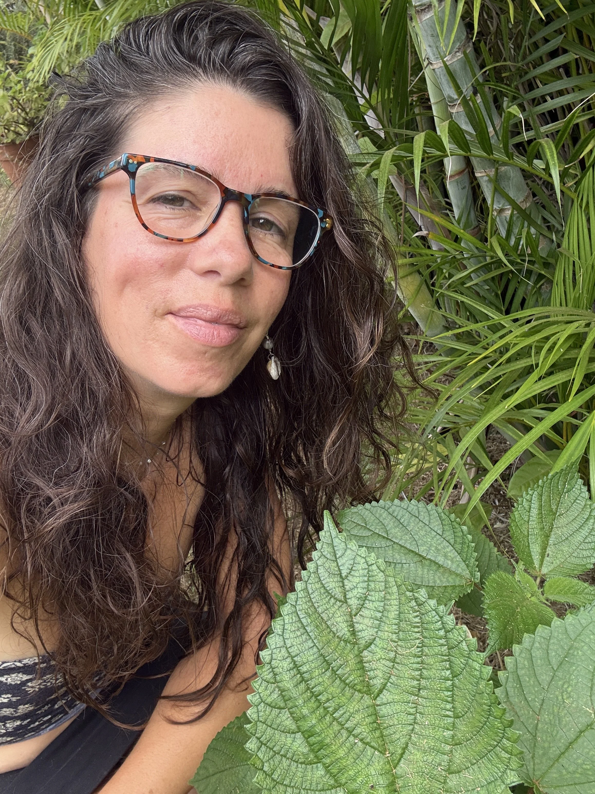 Retrato de una mujer con gafas de montura color tortuga, cabello rizado y largo, y con fondo de plantas verdes tropicales.