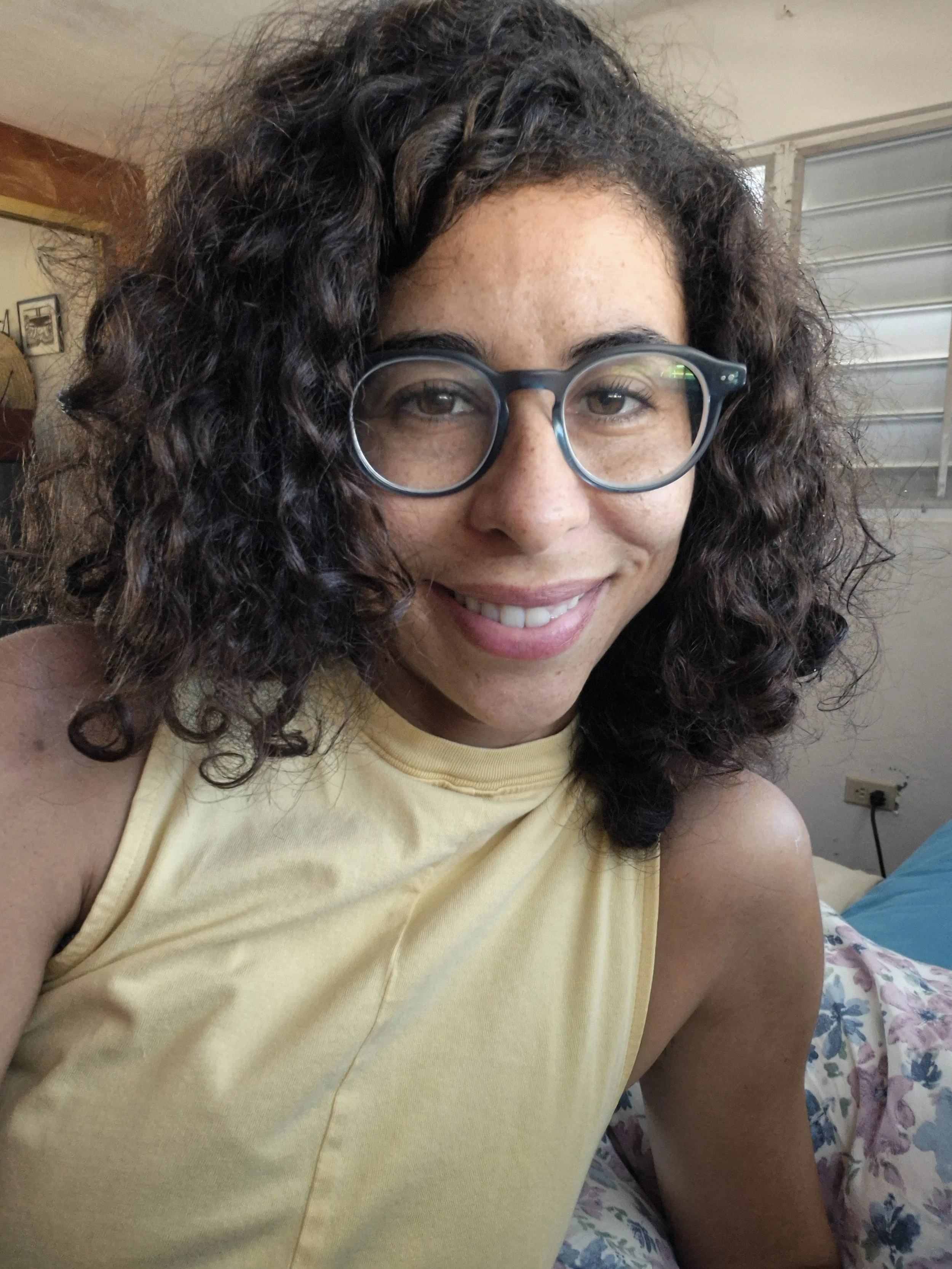 Retrato de una mujer con cabello rizado oscuro, gafas redondas, sonriente, vestida con camiseta amarilla, sentada en una cama con cobijas de flores, en una habitación con paredes claras y ventanas