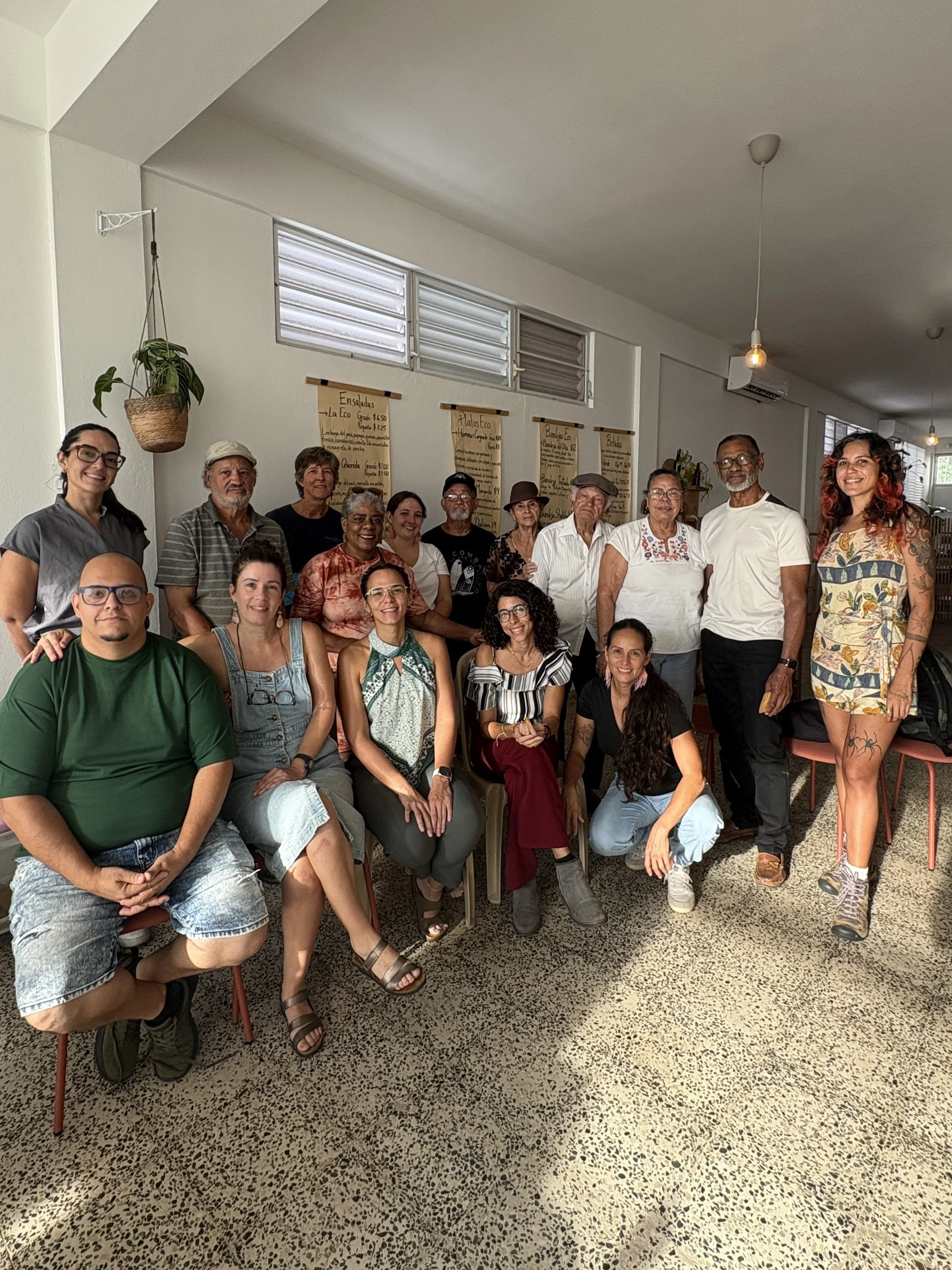 Grupo de personas posando en un espacio interior con paredes blancas y ventanas de ventilación. Algunos están de pie y otros sentados, parecen ser un grupo diverso de adultos en una reunión o evento social.