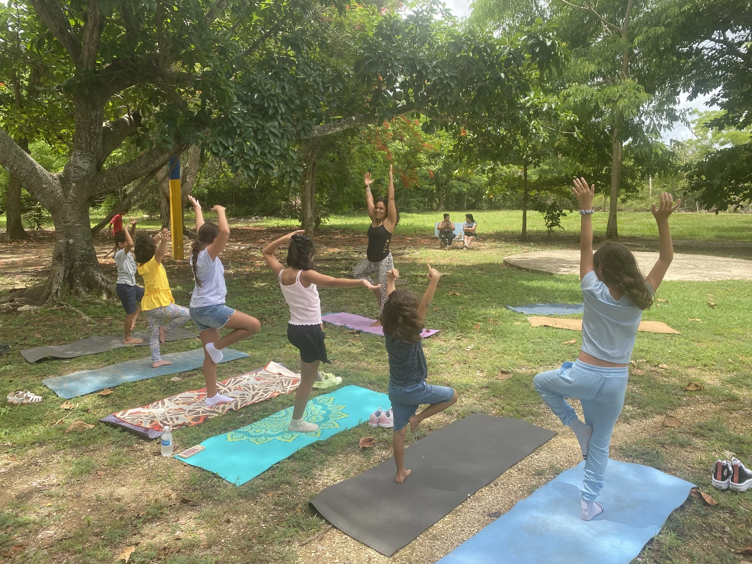 Un grupo de niños y una instructora practicando yoga en un parque con árboles y césped, algunas personas sentadas en bancos en el fondo.