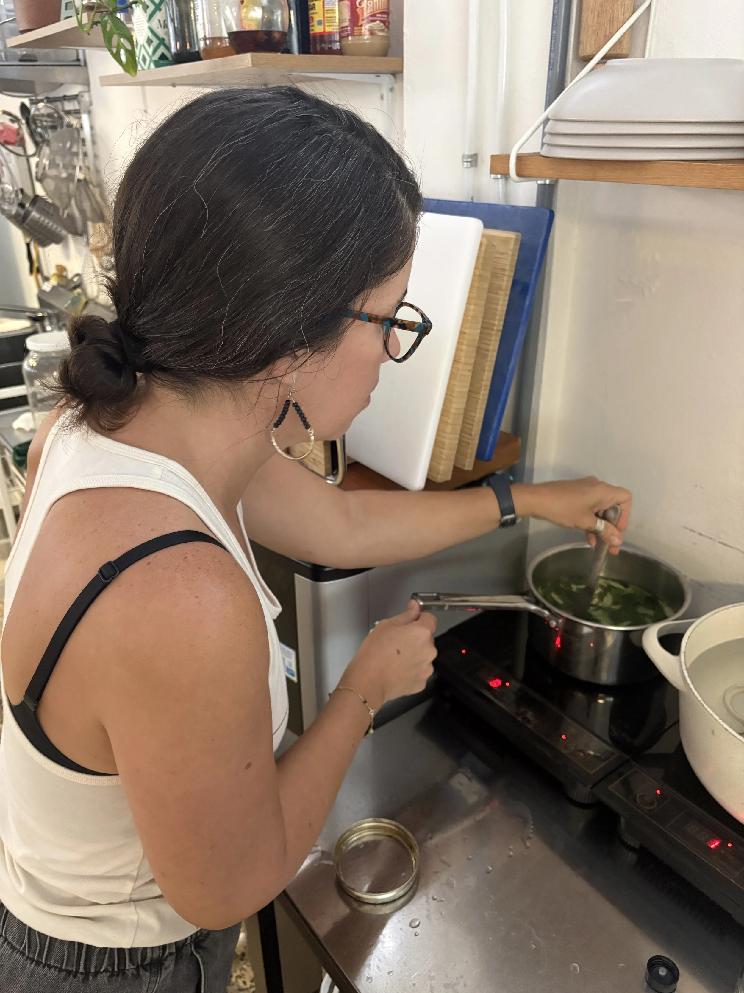 Una mujer con gafas, pendientes y cabello oscuro atada en un chongo, está cocinando en una estufa eléctrica. Ella remueve una olla con sopa o caldo. La cocina tiene varios utensilios y recipientes.