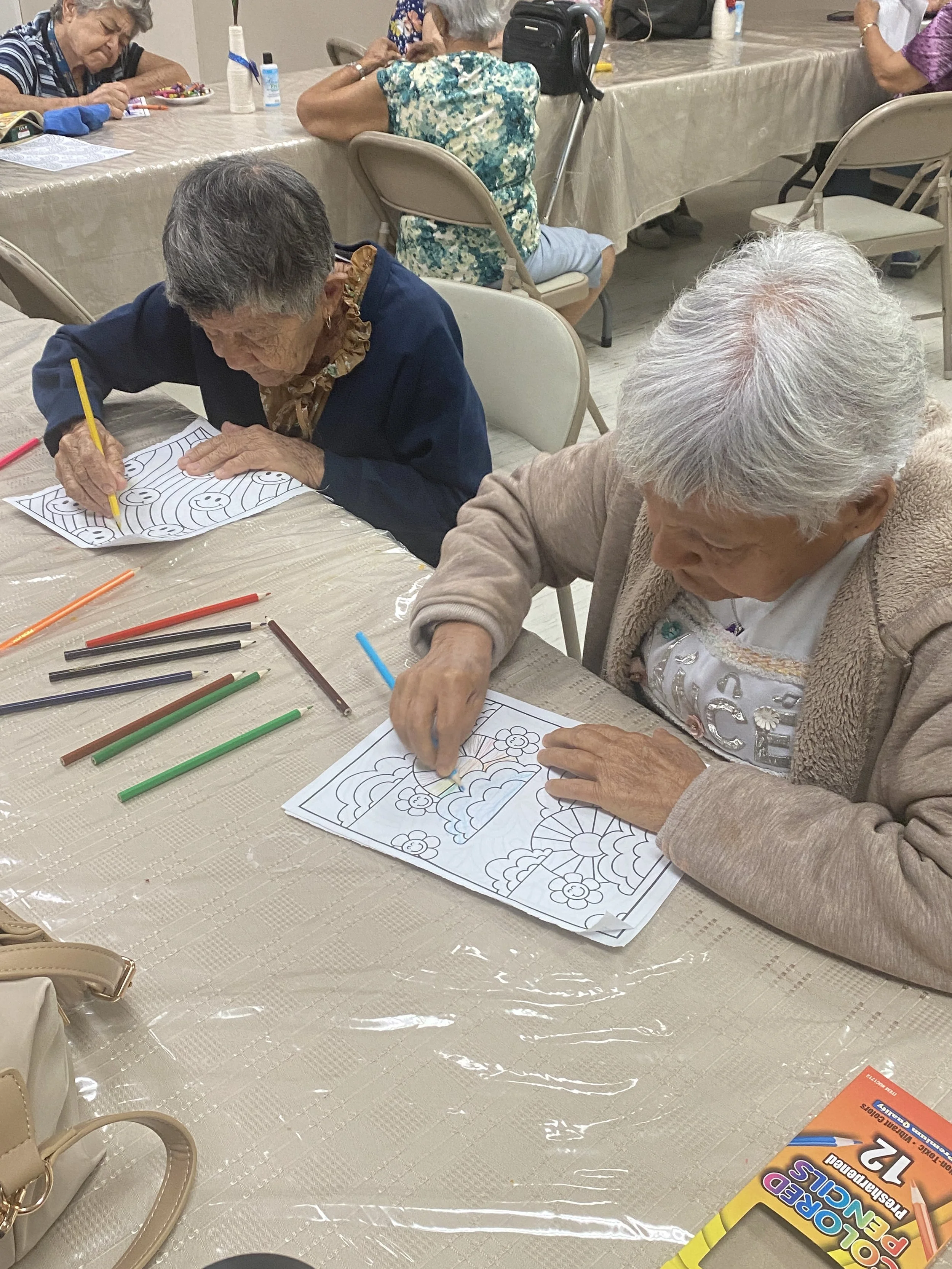 Adultos mayores sentados en una mesa de actividades coloreando dibujos con lápices de colores en un ambiente de recreación o taller.