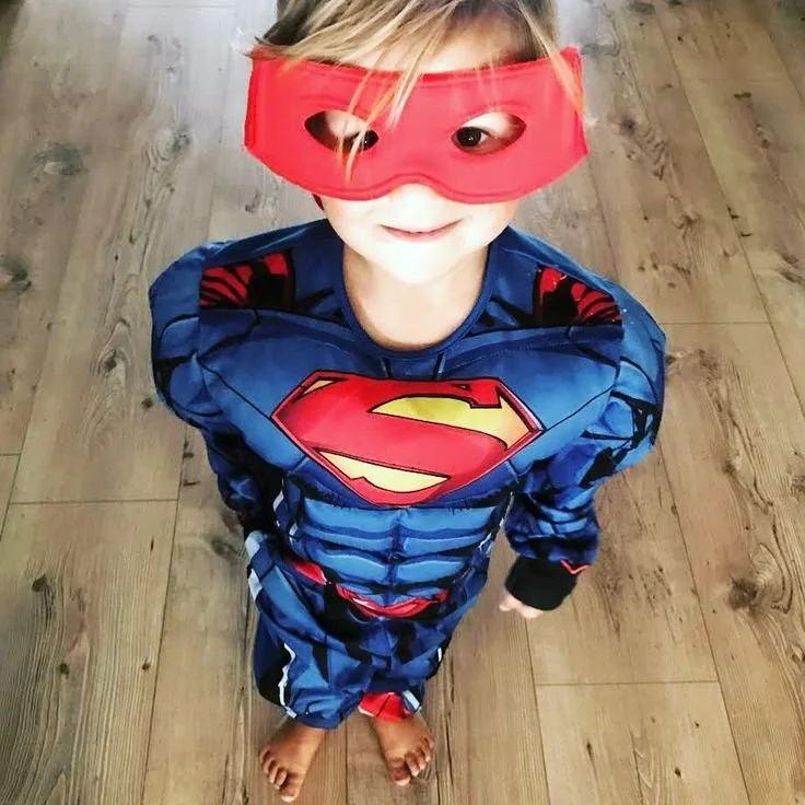 Ein Kind in einem Superman-Kostüm mit einer roten Augenmaske, das barfuß auf einem Holzboden steht.