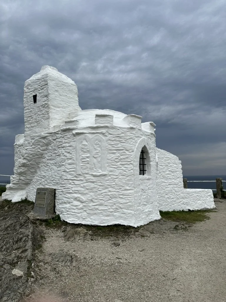 White stone chapel with a tower, small windows, and a dark metal barred window, set against a cloudy sky on a gravel path.