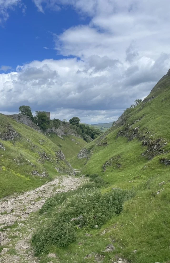 A rocky dirt trail runs through a lush green valley flanked by steep grassy hills under a partly cloudy sky.