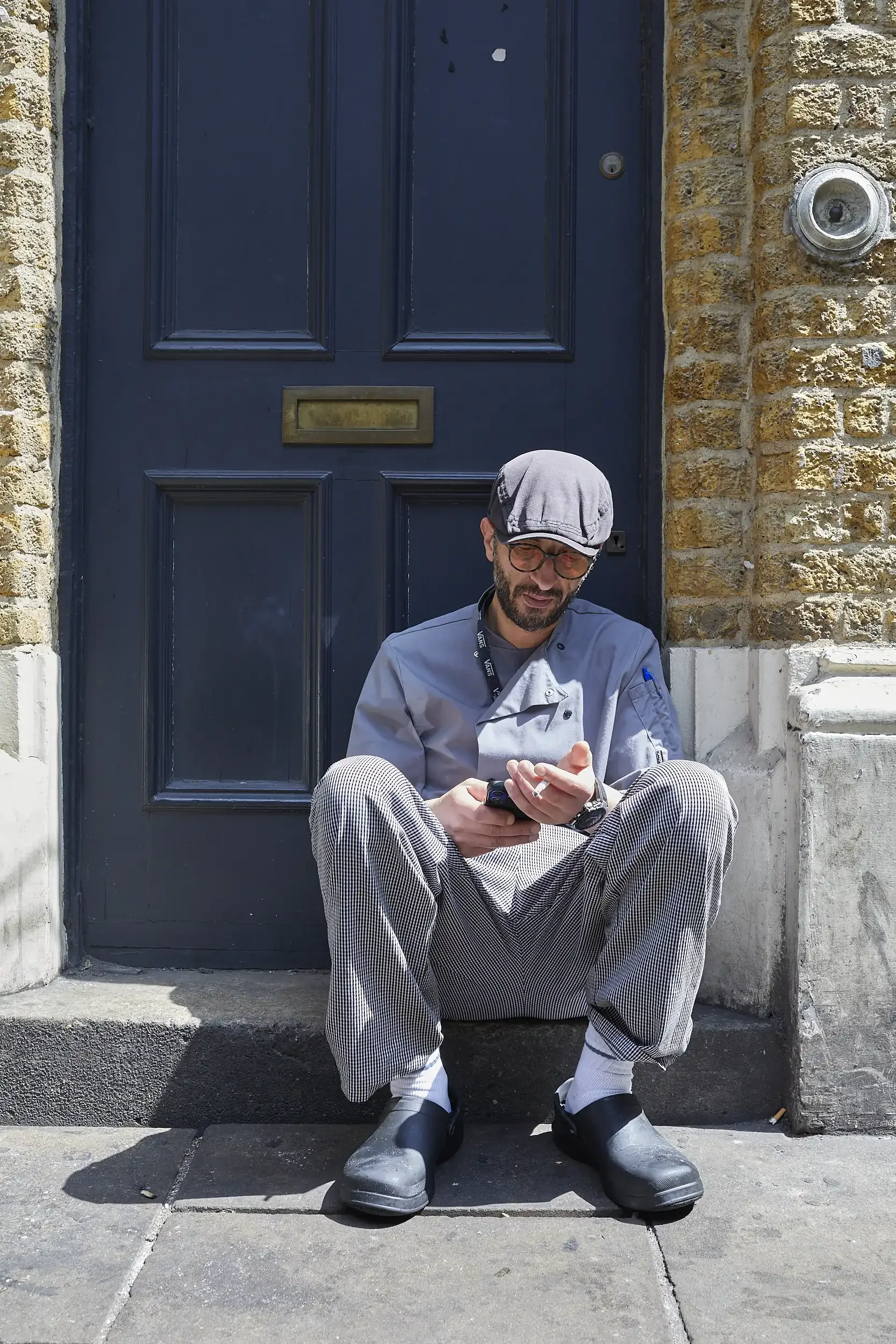 A man with glasses, a beard, and a gray cap sitting on steps outside a brick building, looking at his phone.