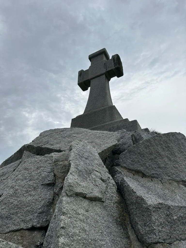 A large stone cross monument at the top of a rocky hill under a cloudy sky.