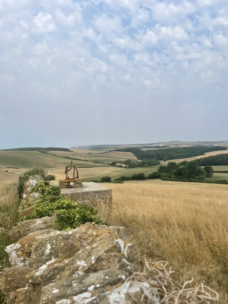 A rural landscape with rolling green hills and fields, a stone wall in the foreground, a small wooden sculpture on a stone topped with stacked stones, under a partly cloudy sky.