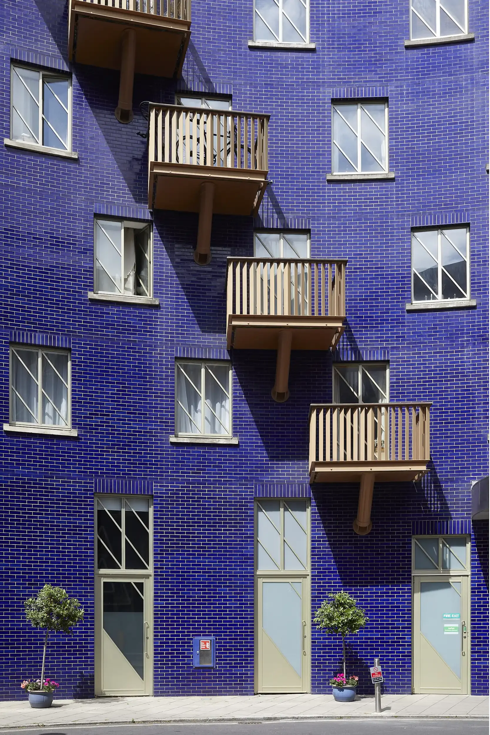 A building with a distinctive blue brick facade featuring several small trees in pots on the sidewalk. There are three balconies with wooden railings and brown supports. The building has multiple rectangular windows and doors, with some signs visible