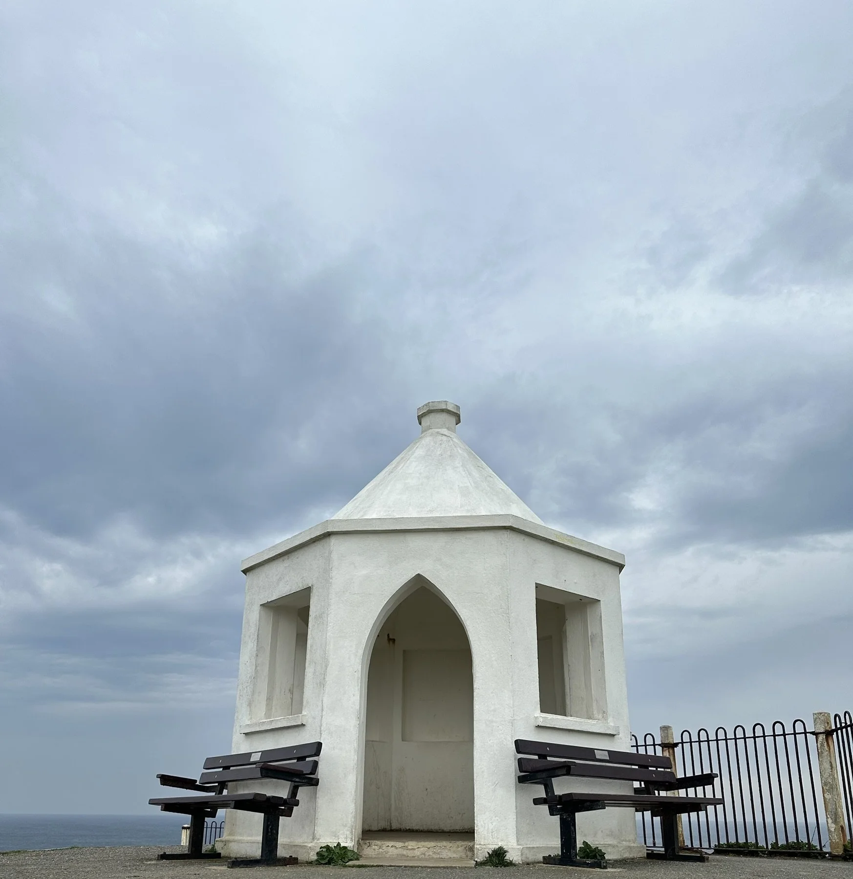 White pavilion structure with arched openings, located outdoors with a view of the water in the background. There are benches on either side of the pavilion and a fence nearby. The sky is cloudy and overcast.