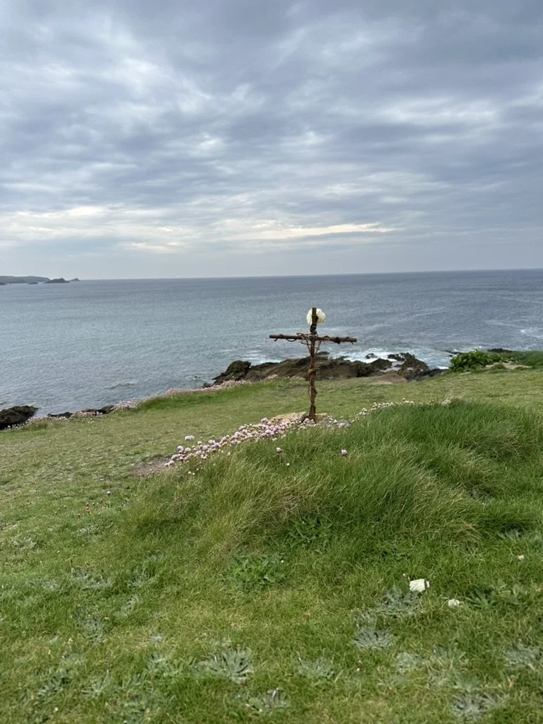 A small island with green grass and pink flowers, overlooking the ocean with rocky shoreline, a weathered cross made of driftwood, and a cloudy sky.