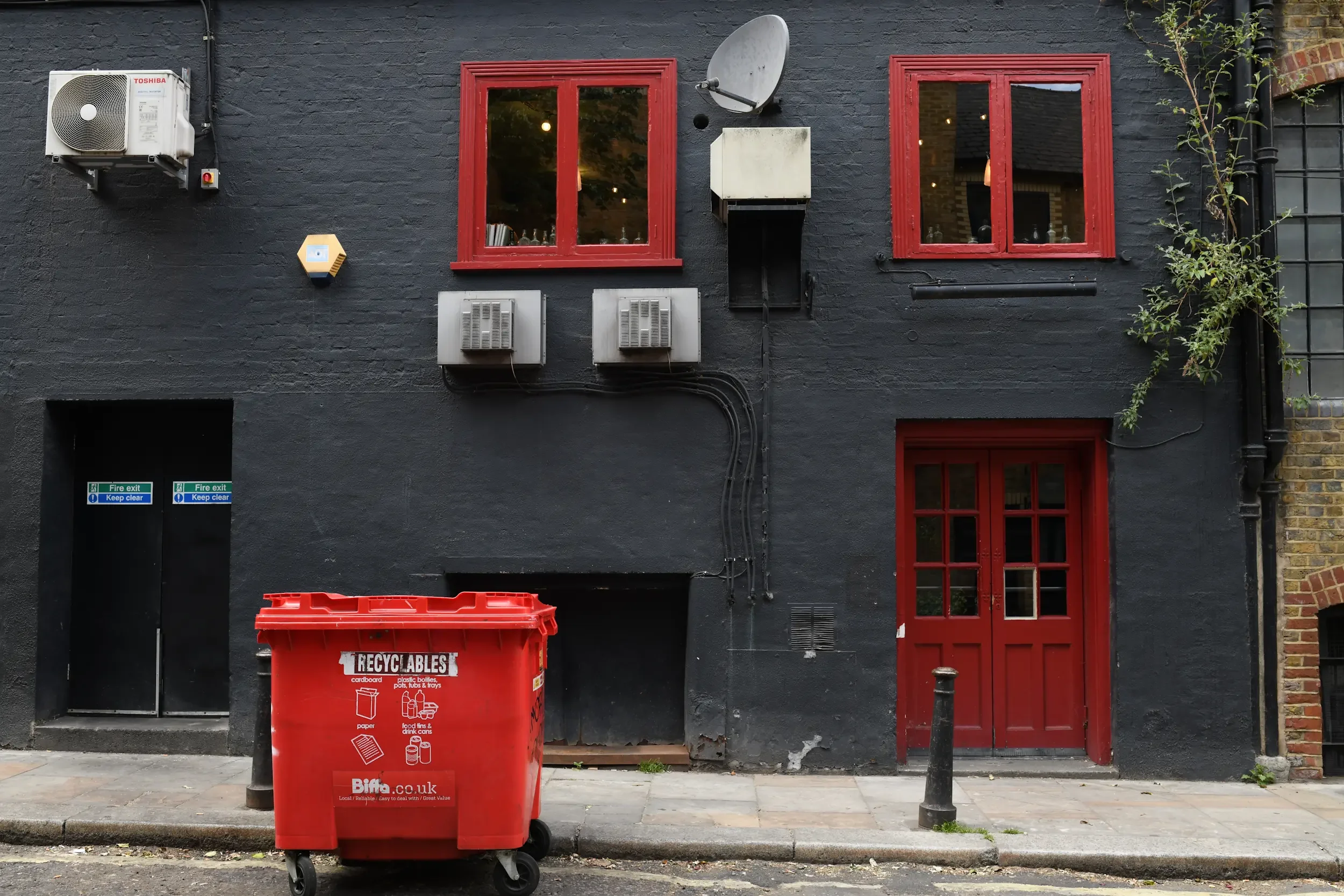 A building with black painted brick wall, red window frames, and a red door. There are two air conditioning units, a satellite dish, and a fire exit sign on the wall. A large red recycling bin is on the sidewalk in front of the building. Some greener